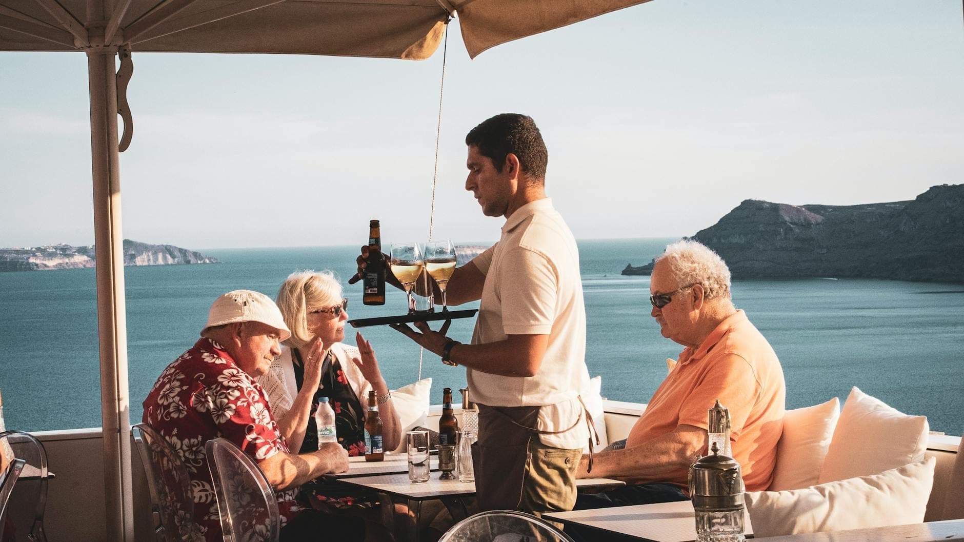 Gäste genießen eine Weinprobe auf einer Klippen-Terrasse mit Blick auf Santorins Caldera – ein Mitarbeiter schenkt Wein ein, im Hintergrund die beeindruckende Vulkanlandschaft.