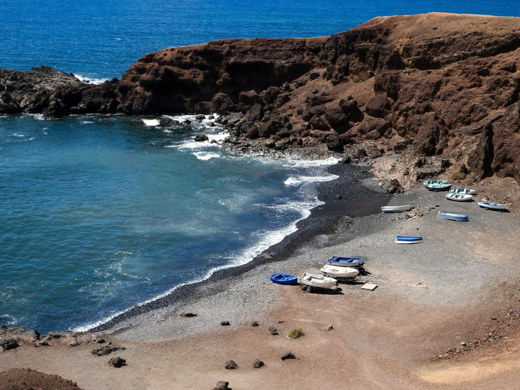 Geschwungener Schwarzsandstrand in Santorini mit kleinen Booten und vulkanischen Klippen rund um die Bucht bei hellem Sonnenlicht.