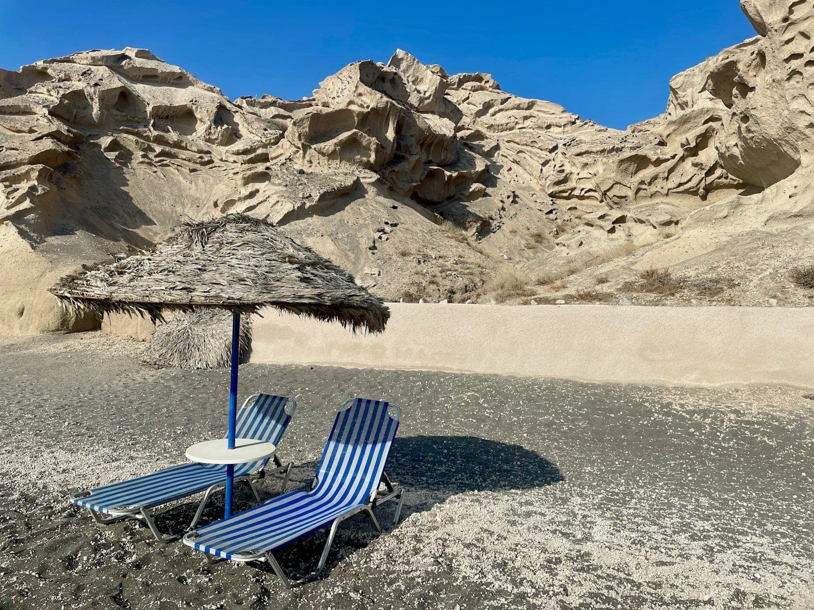 Zwei blau-gestreifte Liegestühle und ein Strohschirm auf einem schwarzen Sandstrand in Santorini, im Hintergrund dramatische Vulkanfelsen unter einem strahlend blauen Himmel.