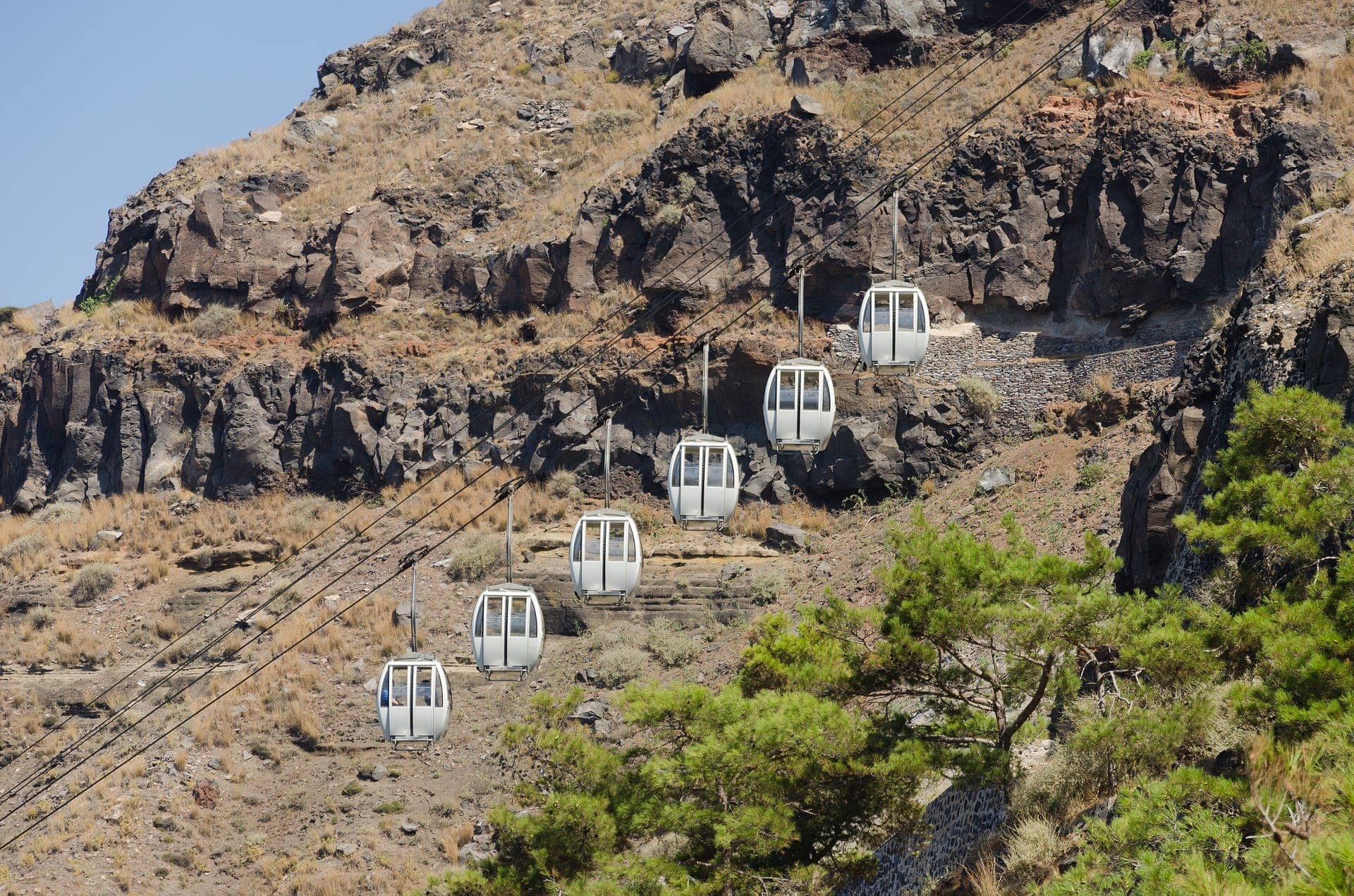 Die Seilbahngondeln von Santorini fahren an einer felsigen, sonnenbeschienenen Steilküste empor, die mit trockenem Gras und spärlichem Grün unter einem klaren blauen Himmel gesprenkelt ist.