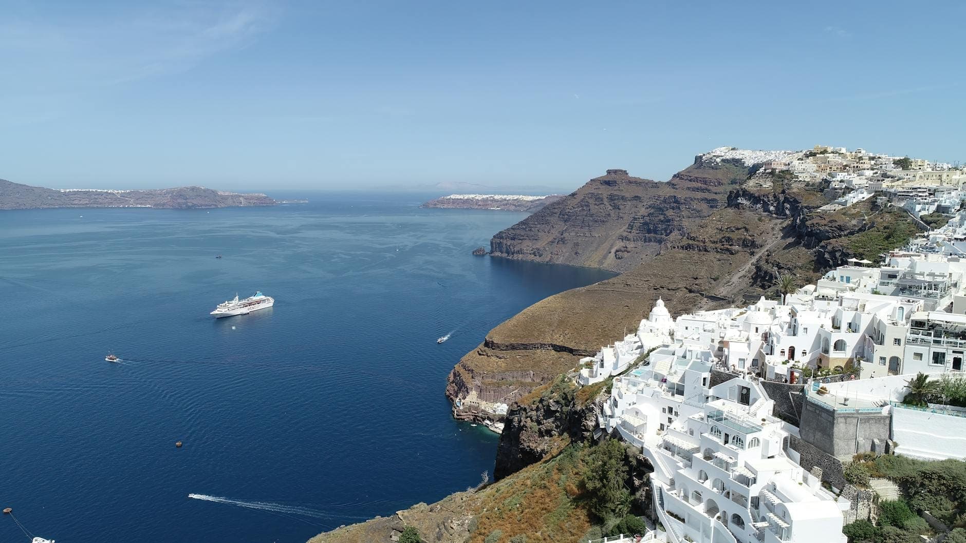 Dramatische Luftaufnahme der Santorin Caldera mit steilen Klippen, leuchtendem blauem Meer, einem weißen Kreuzfahrtschiff und typischen weiß gekalkten Häusern am Kraterrand.