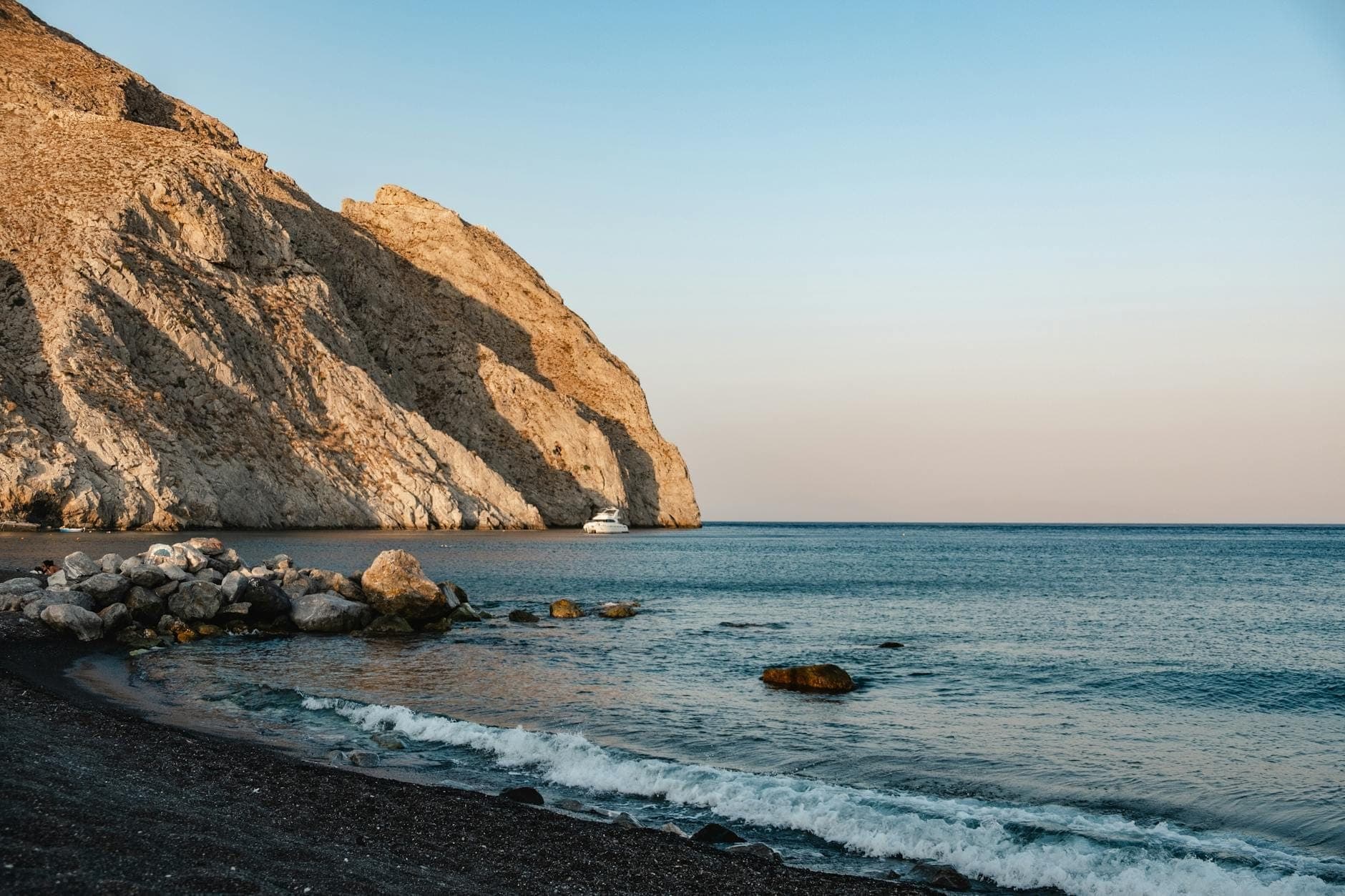 Una tranquila y salvaje playa de Santorini al atardecer con arena negra, costa rocosa, acantilados y suaves olas bajo un cielo despejado.