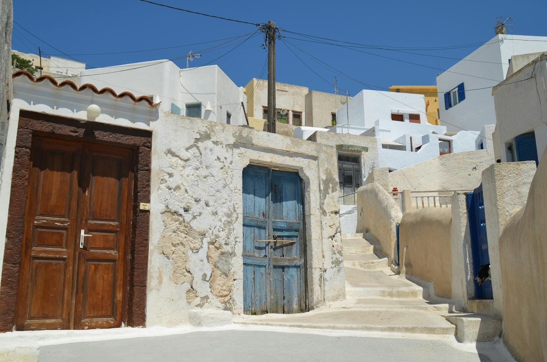 Casas de piedra desgastadas con puertas de madera rústicas y escaleras encaladas sinuosas en un pueblo tradicional de Santorini bajo un cielo azul.