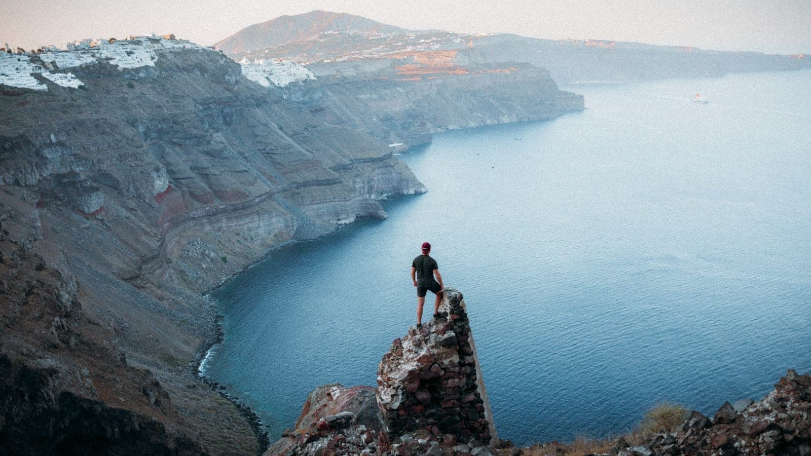 Ein Wanderer steht auf einem felsigen Vorsprung mit Blick auf die dramatischen Caldera-Klippen Santorins und das tiefblaue Meer beim Sonnenuntergang – Abenteuer und atemberaubende Insellandschaft pur.