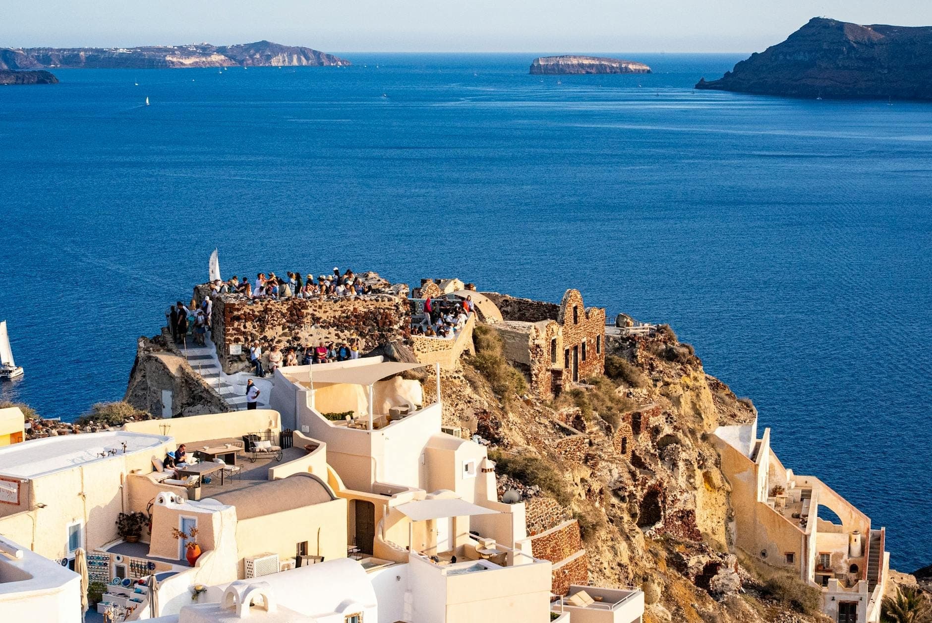 Blick auf die Ruinen der Burg von Oia mit Menschenmassen auf einem Felsen über dem blauen Meer, weiß getünchte Häuser unter klarem Himmel auf Santorini.