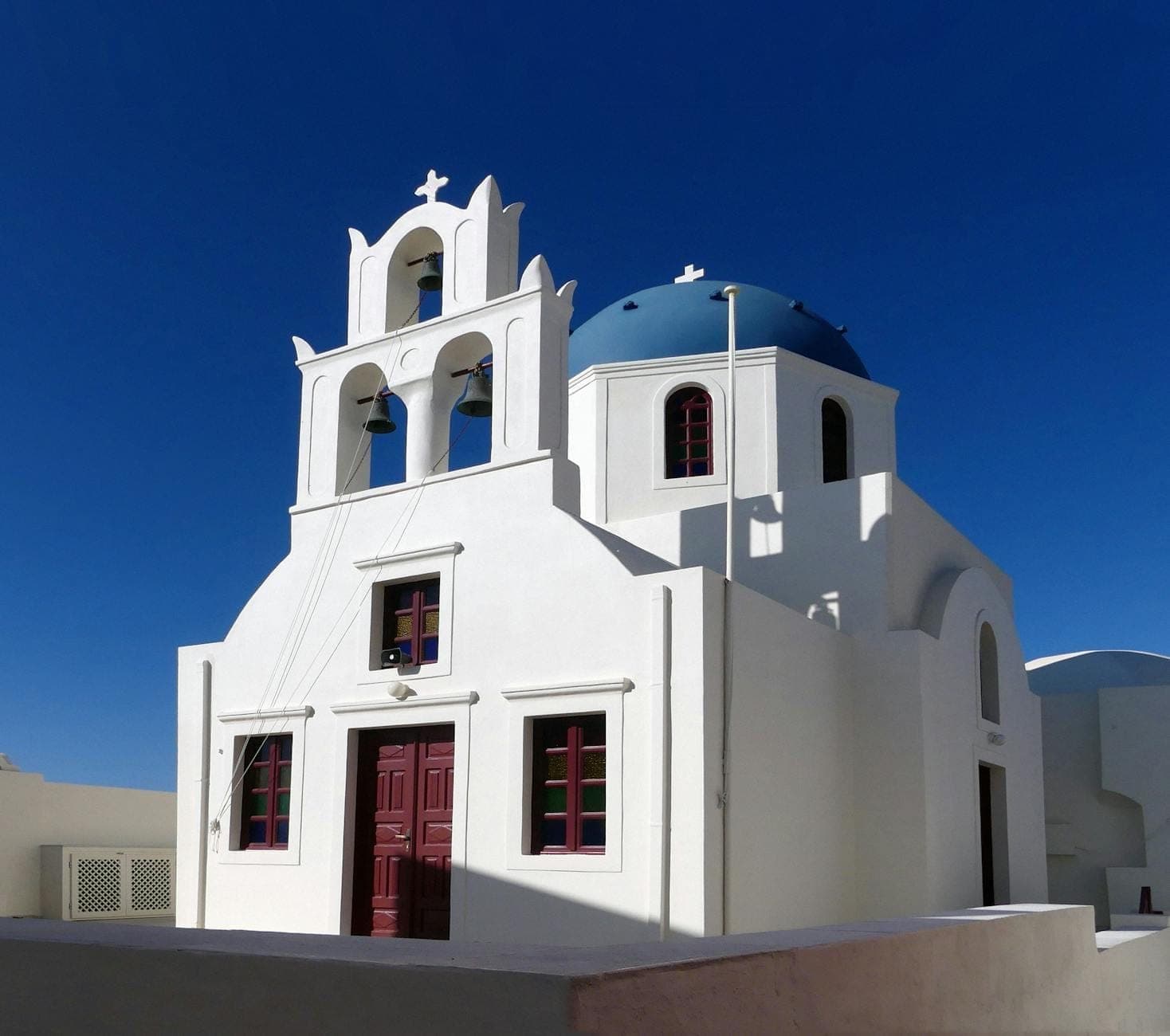 Weiß getünchte Kirche mit blauer Kuppel, drei Glockentürmen und roten Türen vor einem tiefblauen Himmel in Santorini.