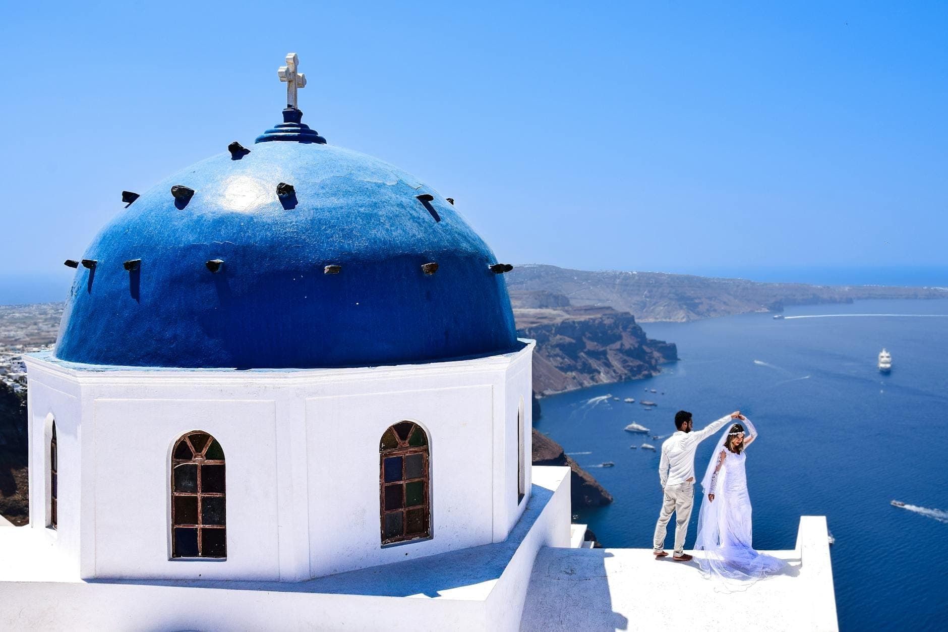 Brautpaar umarmt sich auf einer Terrasse neben Santorinis ikonischer Blaukuppelkirche, mit beeindruckenden Caldera-Klippen und tiefblauem Meer unter klarem Himmel.