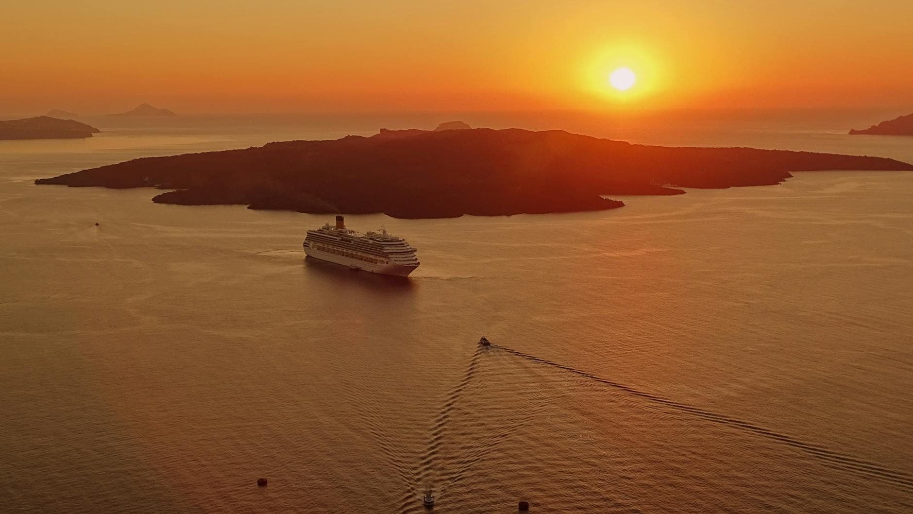Grande nave da crociera che naviga nella caldera di Santorini al tramonto con l'isola vulcanica sagomata contro un cielo arancione.