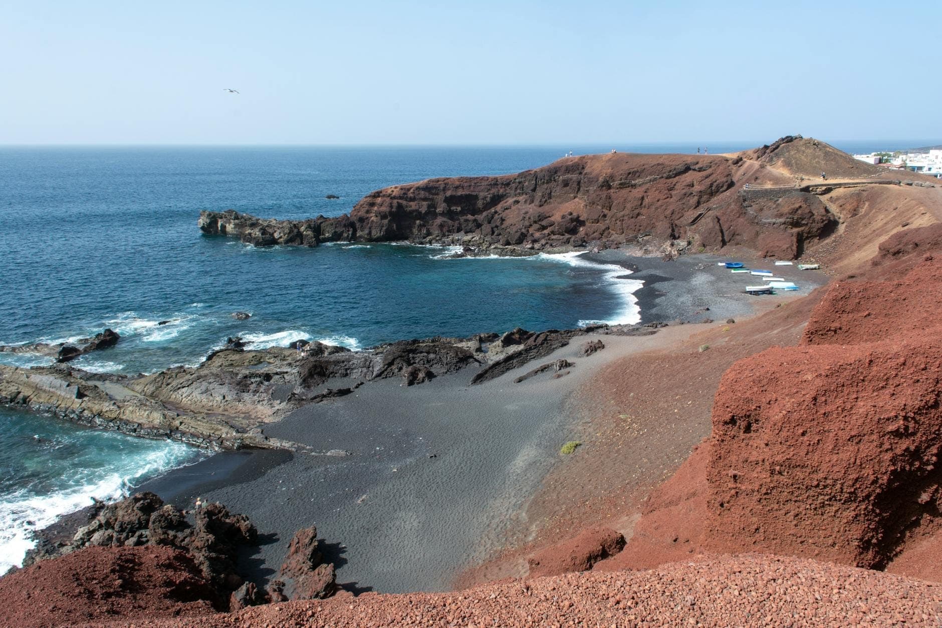 Costa di Santorini con scogliere rosso ruggine e spiaggia di sabbia vulcanica scura che si incontra con il mare blu sotto un cielo limpido.
