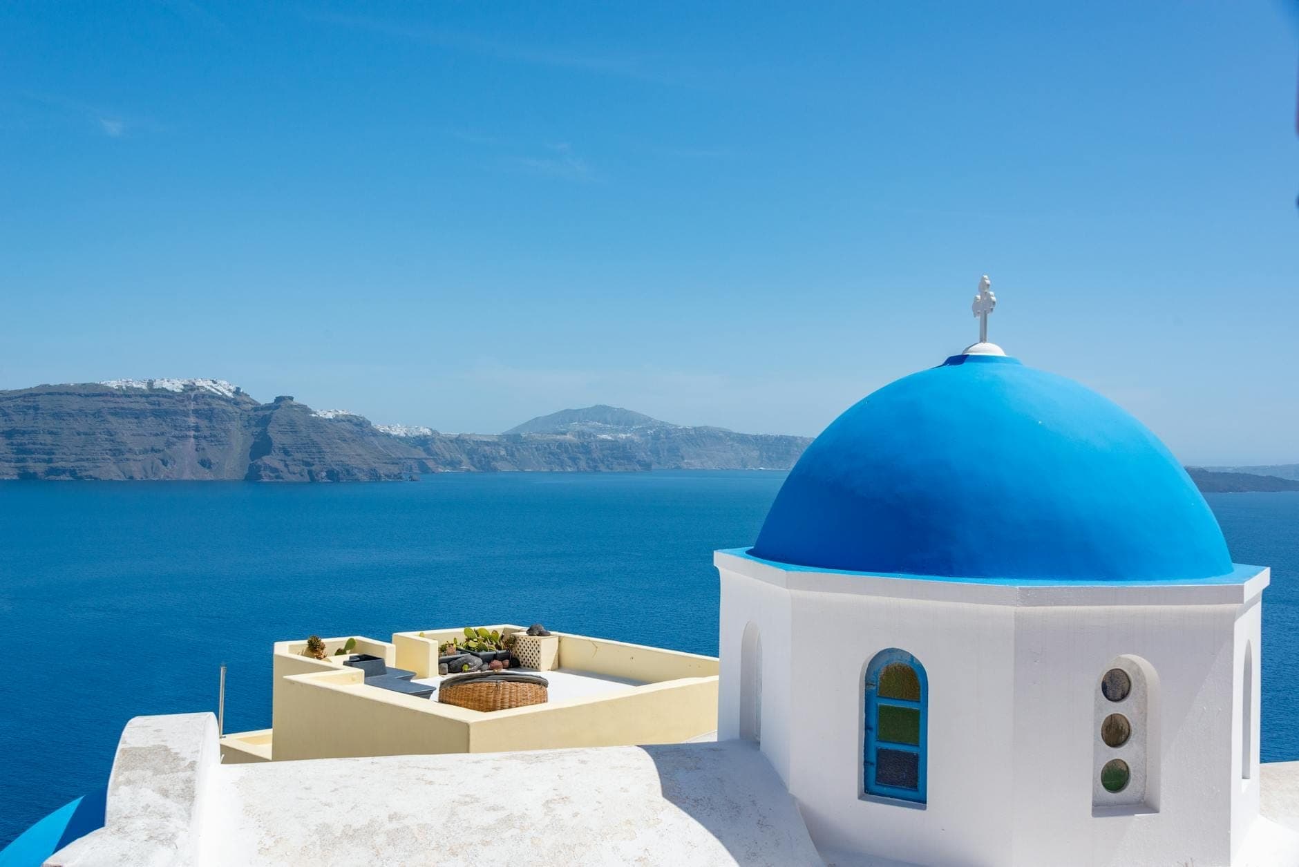 Klassisches Santorin-Panorama mit einer leuchtend blaubedachten Kirche mit Blick auf das tiefblaue Ägäische Meer, vulkanische Klippen im Hintergrund unter klarem Himmel.