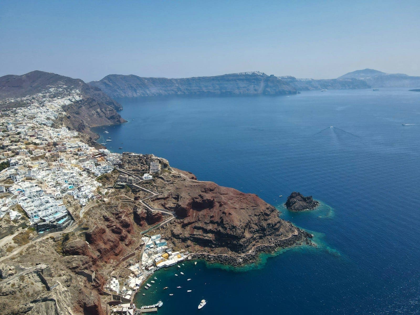 Vista aérea de los dramáticos acantilados de Santorini, edificios encalados y la caldera de intenso azul con yates de lujo anclados frente a la costa en un día soleado.
