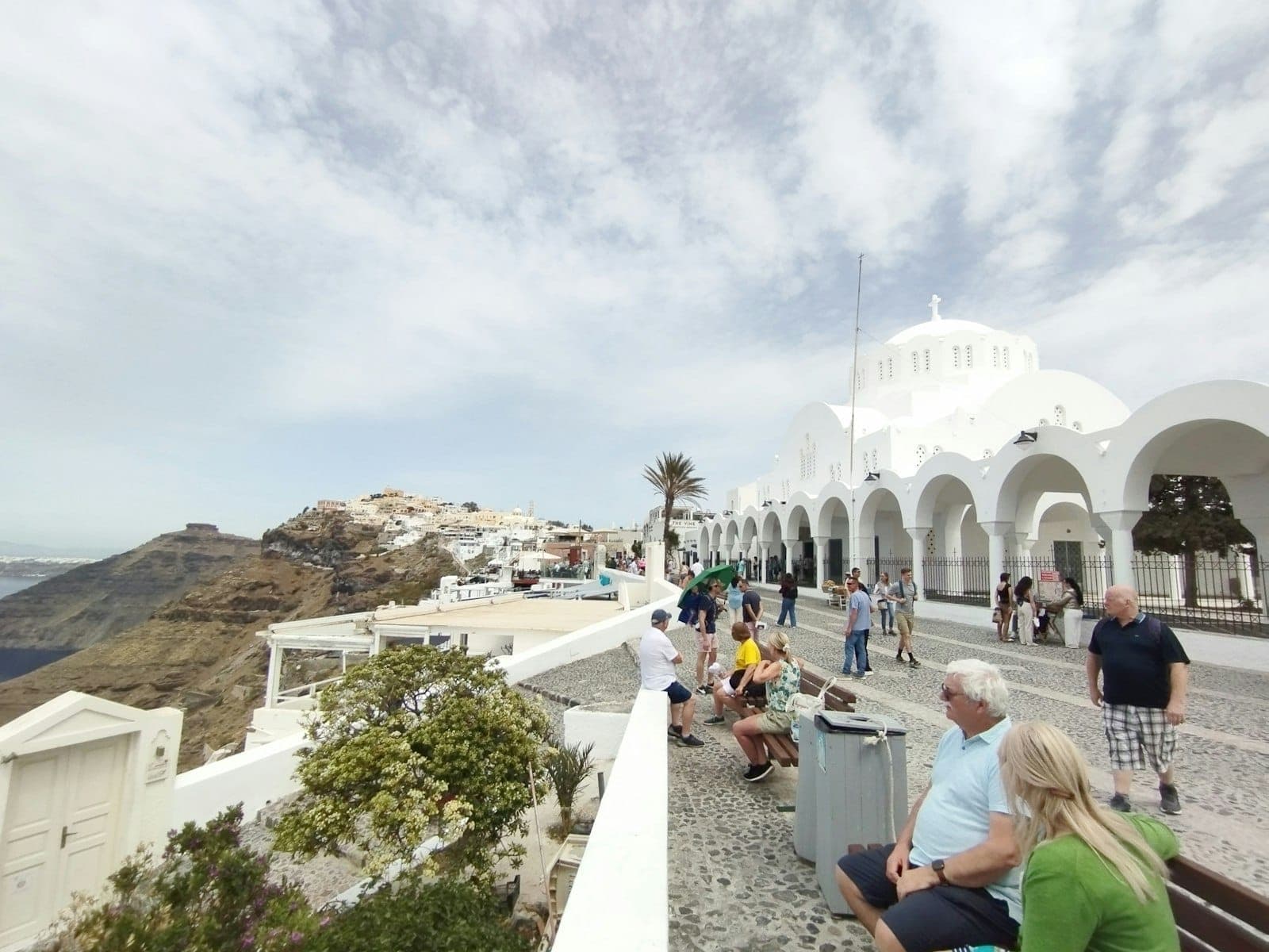 Personas sentadas y caminando cerca de los edificios de arcos blancos de Fira, con una vista lejana de la caldera de Santorini y su pintoresca costa.