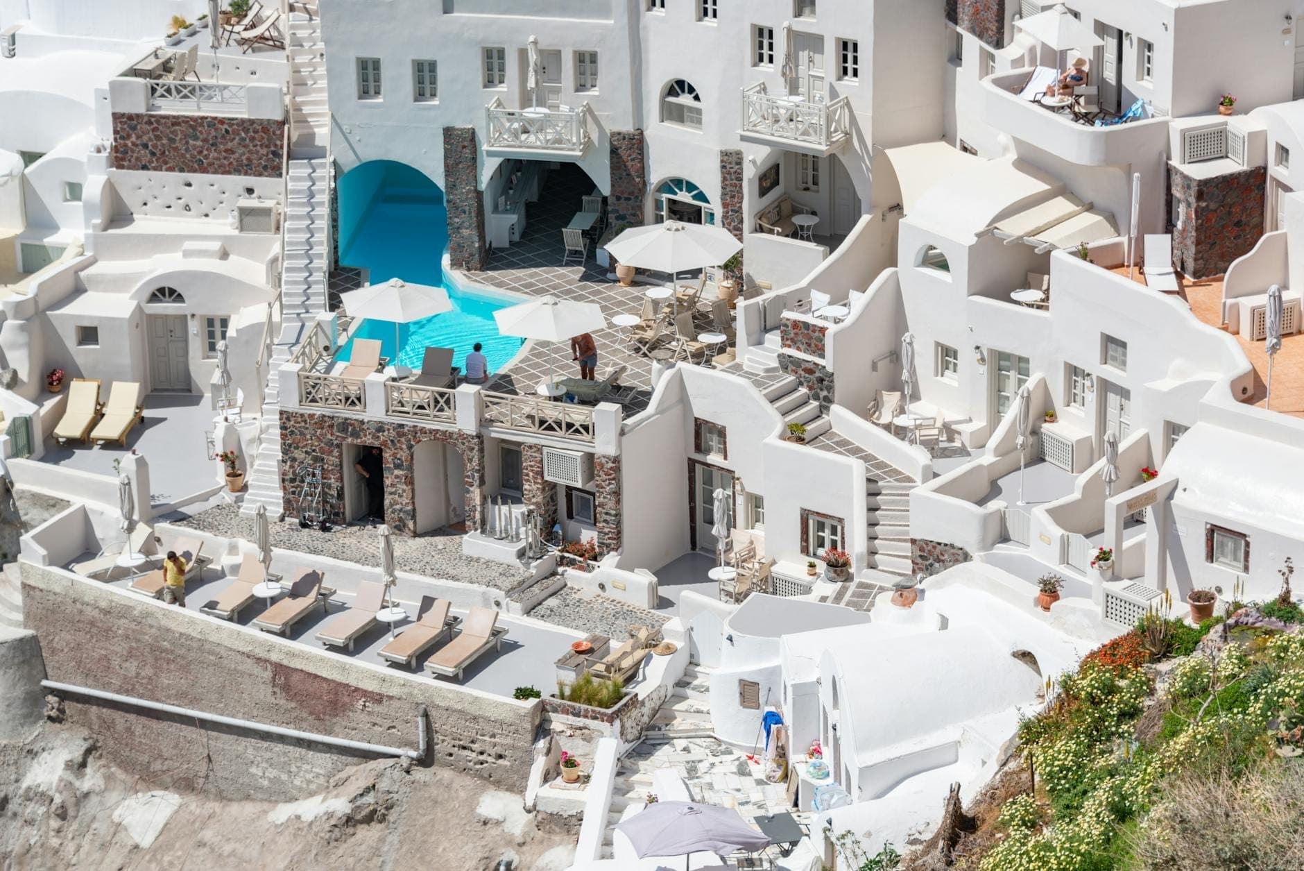 A cluster of classic Santorini white-washed accommodations with balconies, lounge chairs, and a pool, built into the cliffside under bright sunlight.