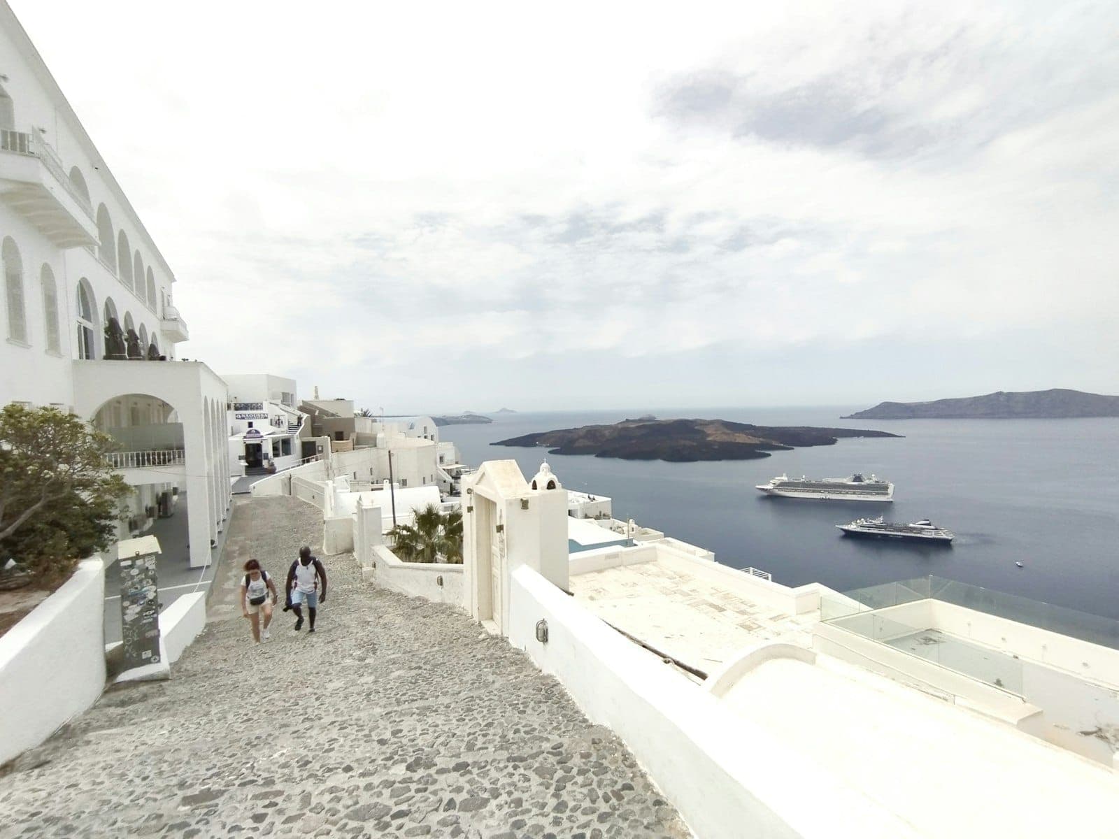People walking along a cobblestone path in Santorini with caldera and cruise ships in the background.
