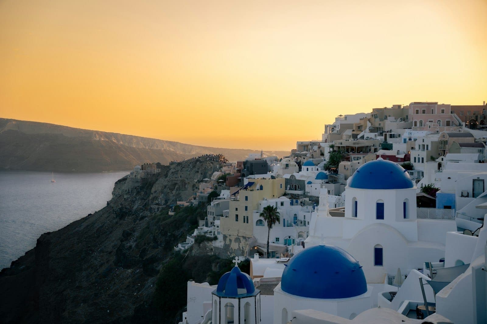 Kalderarand von Santorini mit den ikonischen Kirchen mit blauen Kuppeln, weiß getünchten Gebäuden auf Klippen und einem goldenen Abendhimmel über dem Ägäischen Meer.