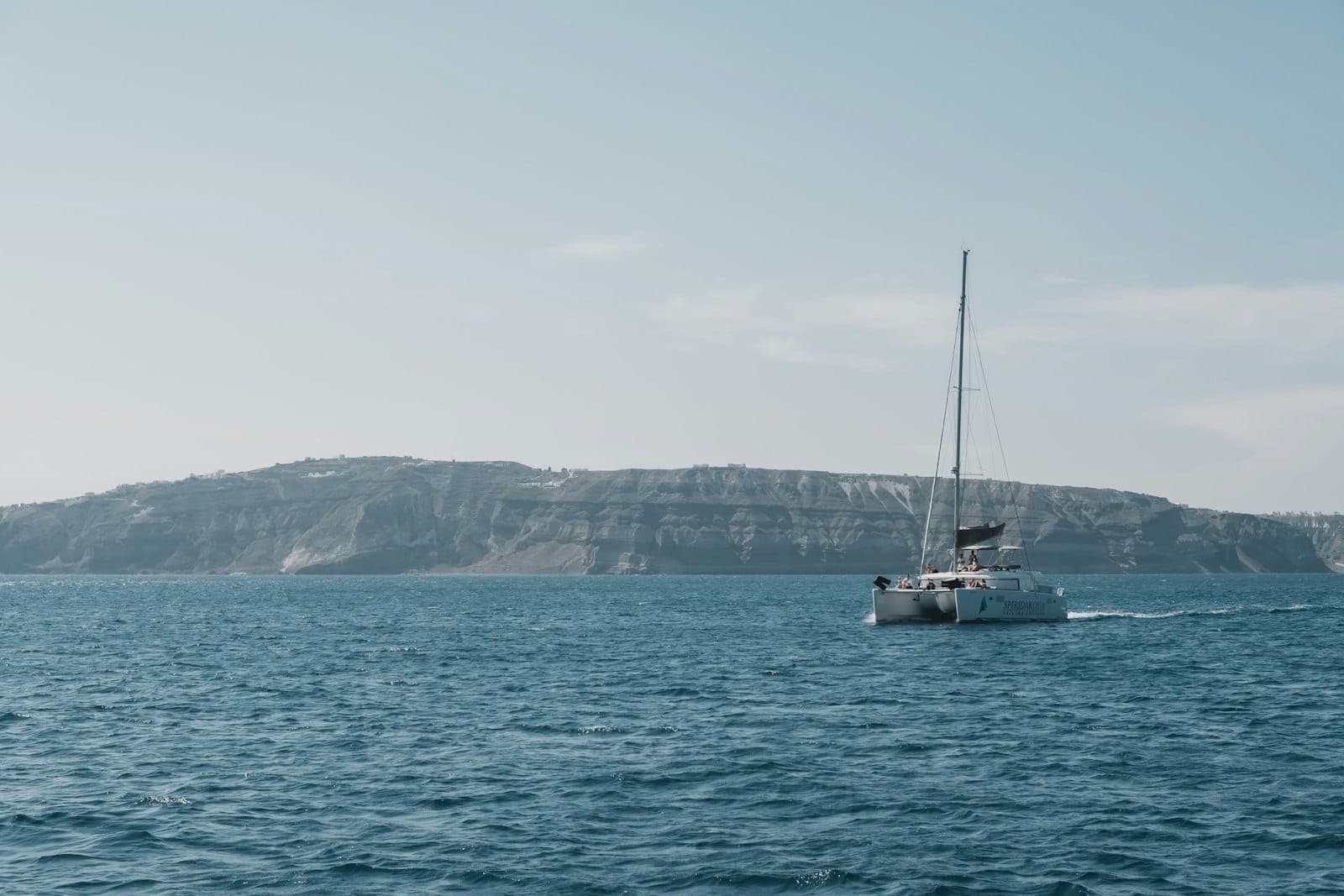 Catamarán navegando cerca de los característicos acantilados de Santorini bajo un cielo despejado, con la costa de la isla al fondo.