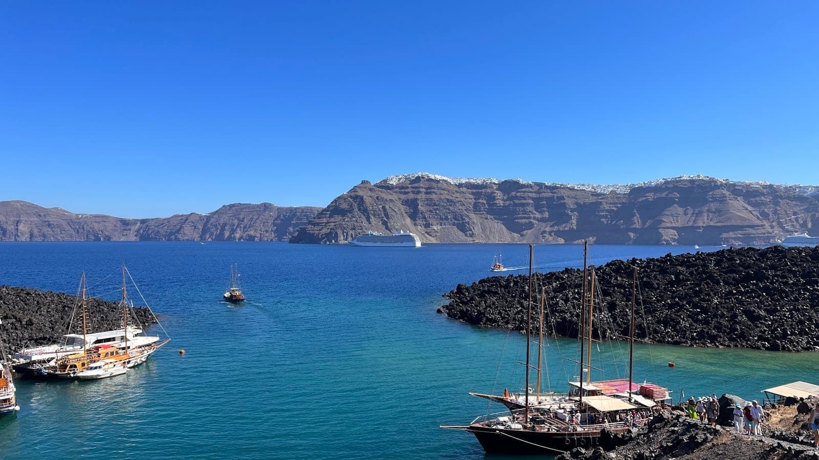 Marina de la caldera de Santorini con veleros tradicionales, personas embarcando y acantilados volcánicos al fondo bajo un cielo azul despejado.
