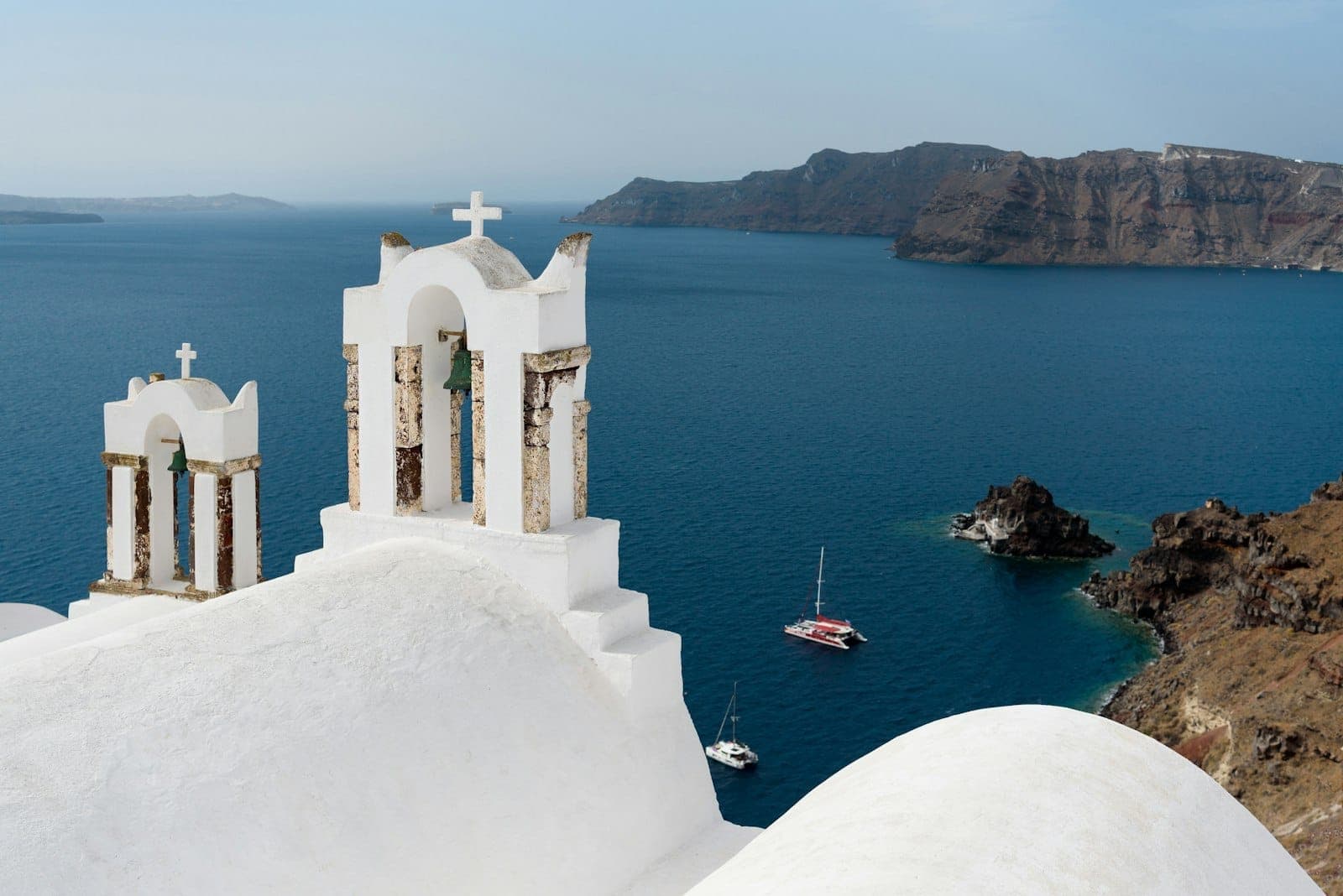 Weiße Kirchenglocken auf einem Dach mit Blick auf das blaue Ägäische Meer, darunter segelnde Katamarane und die Caldera-Klippen Santorinis im Hintergrund.