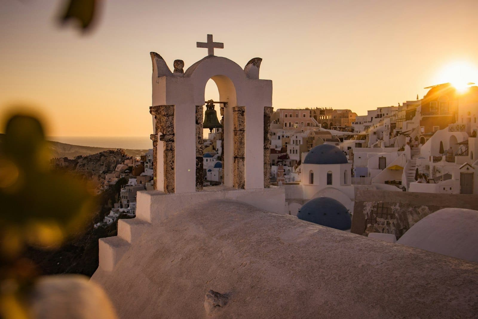 Santorini at sunset with a church bell tower in the foreground, blue domes, whitewashed buildings, and golden sunlight over the iconic clifftop village.