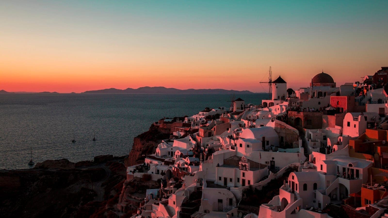 Santorins ikonische weiße Häuser mit blauen Kuppeln über dem Meer bei Sonnenuntergang – mit leuchtendem Orange und Pink am Himmel und der Silhouette einer klassischen Windmühle.