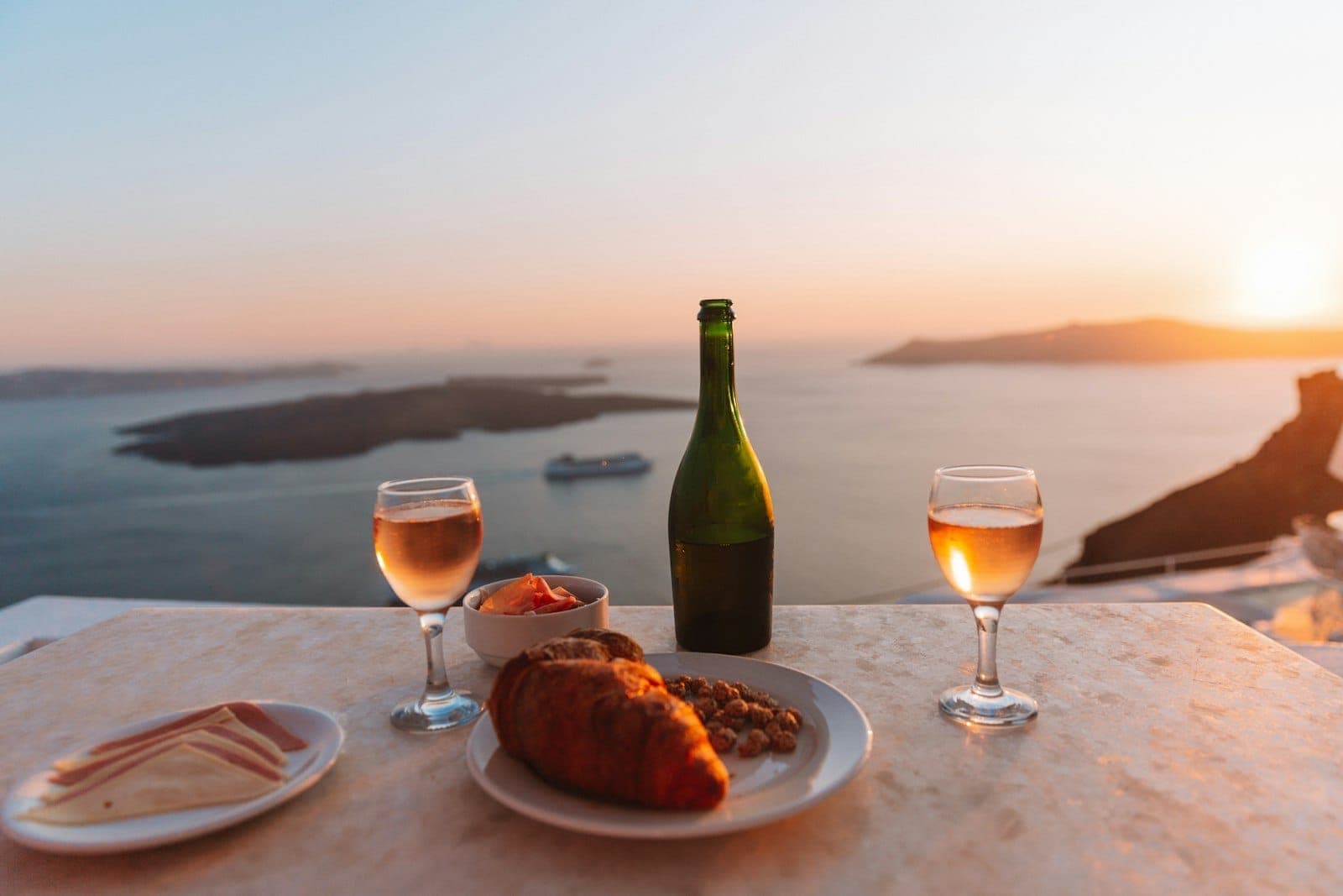 Ein gedeckter Tisch mit Wein, Gläsern und Speisen mit Blick auf die Santorini-Caldera bei Sonnenuntergang, mit goldenem Licht auf dem Meer und den Klippen.