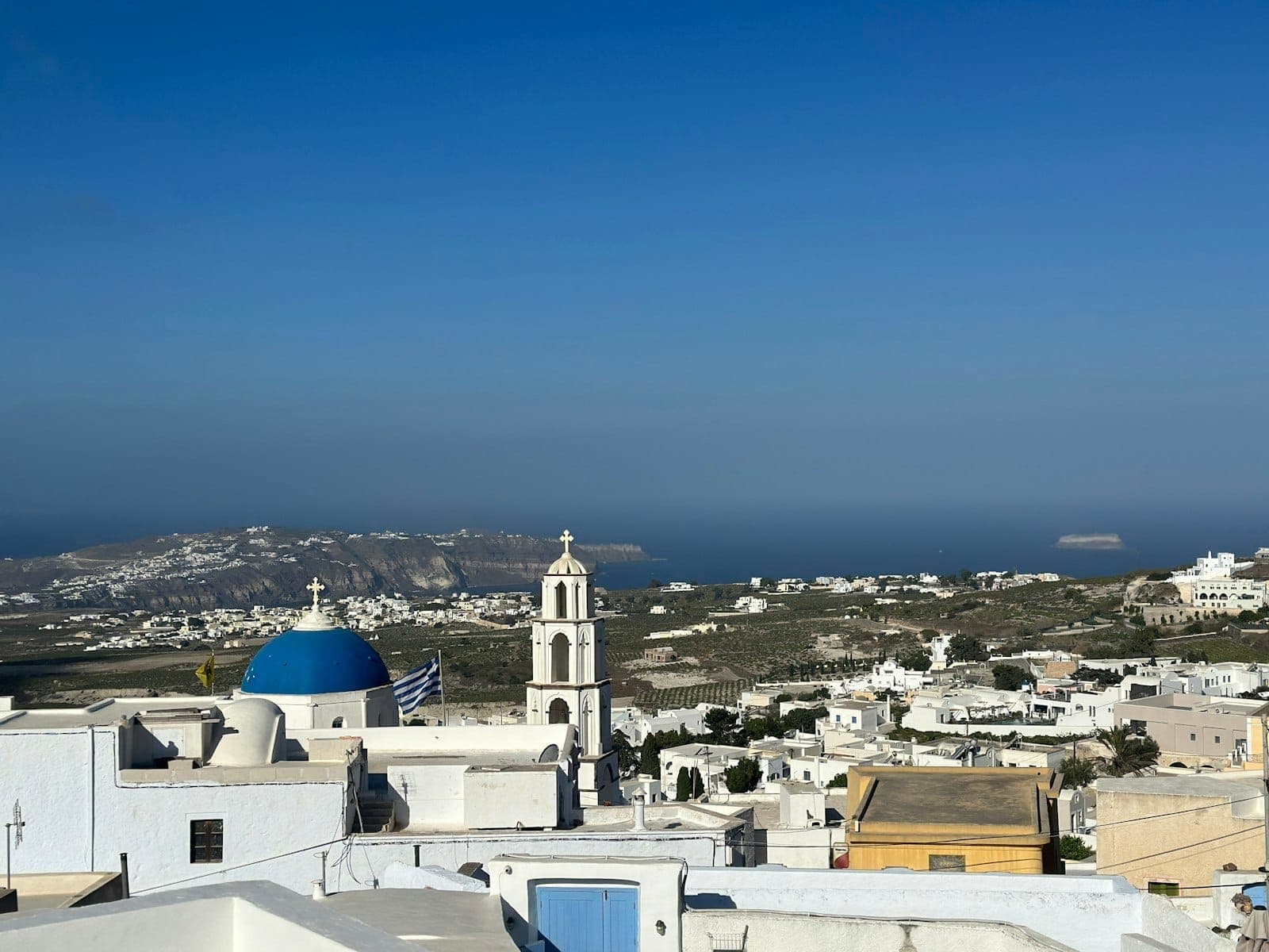 Vista panoramica da un villaggio dell'entroterra di Santorini con edifici bianchi, una chiesa con cupola blu e ampie vedute sull'isola e sul mare.