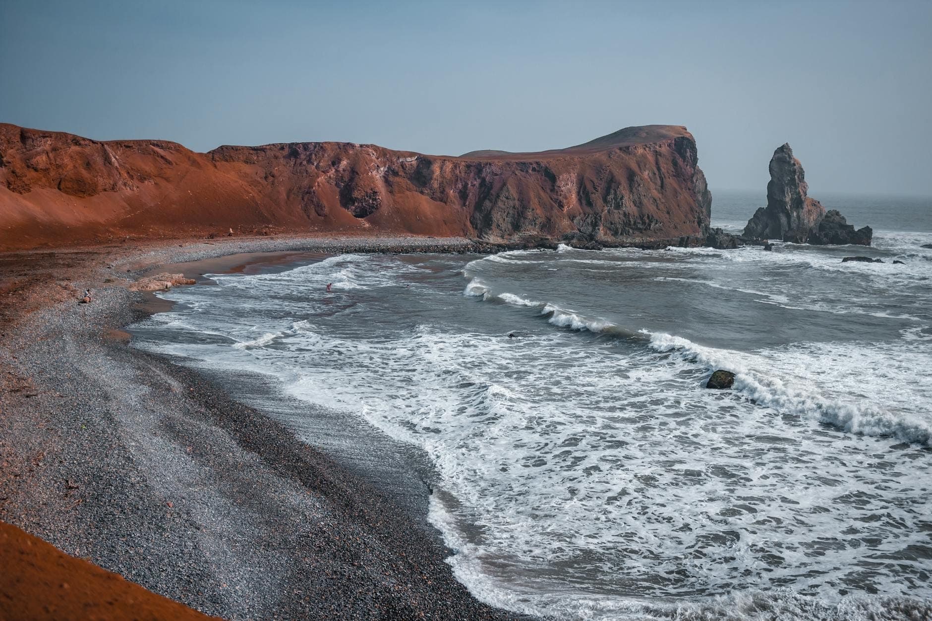 Vista panoramica di una drammatica costa con scogliere rosse, onde che si infrangono su una spiaggia sassosa, un promontorio e uno stack marino a Santorini.