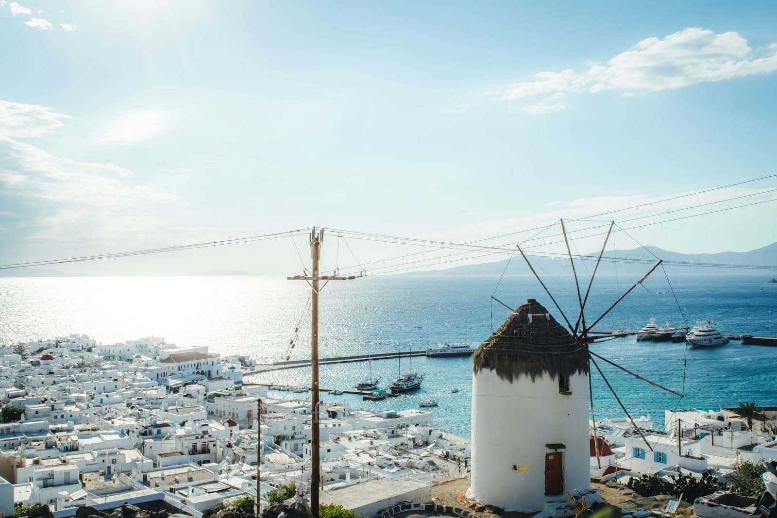 Blick auf die Windmühle von Mykonos mit dem blauen Meer, weiß getünchten Gebäuden und Yachten im Hafen unter strahlend blauem Himmel.