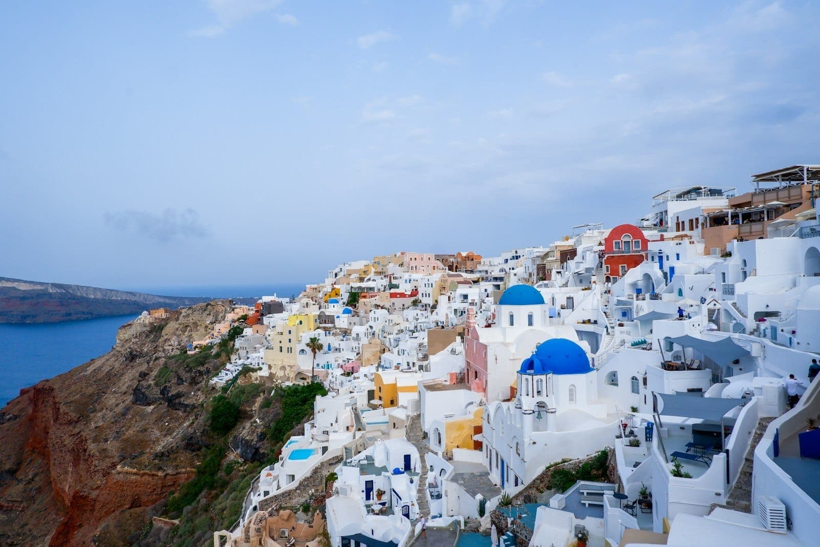 Weiter Blick über Santorinis ikonische weiß getünchte Häuser mit blauen Kuppeln, die Ägäis in tiefem Blau und dramatische Felsenklippen unter einem teils bewölkten Himmel.