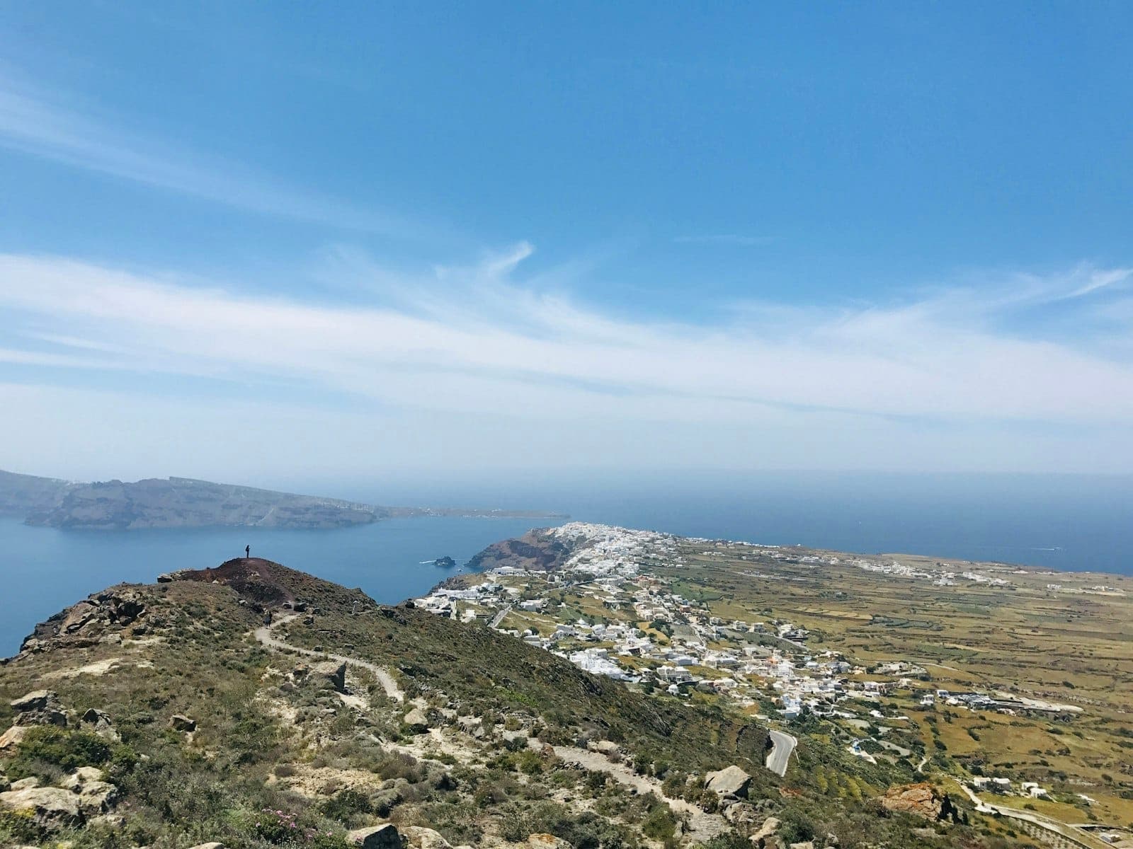 Vista amplia de la caldera de Santorini con cielo despejado, mar, sendero escarpado y lejanos pueblos encalados con vistas a la costa.