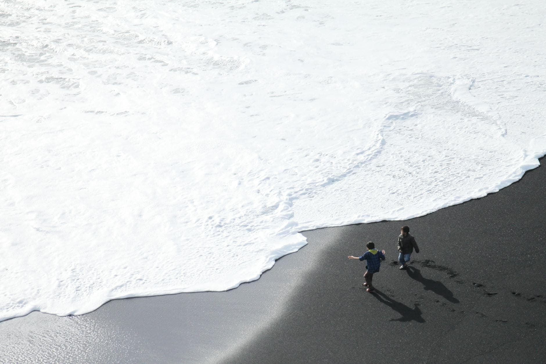 Dos niños caminan por la orilla de las olas en una playa de arena negra bajo la luz del día.