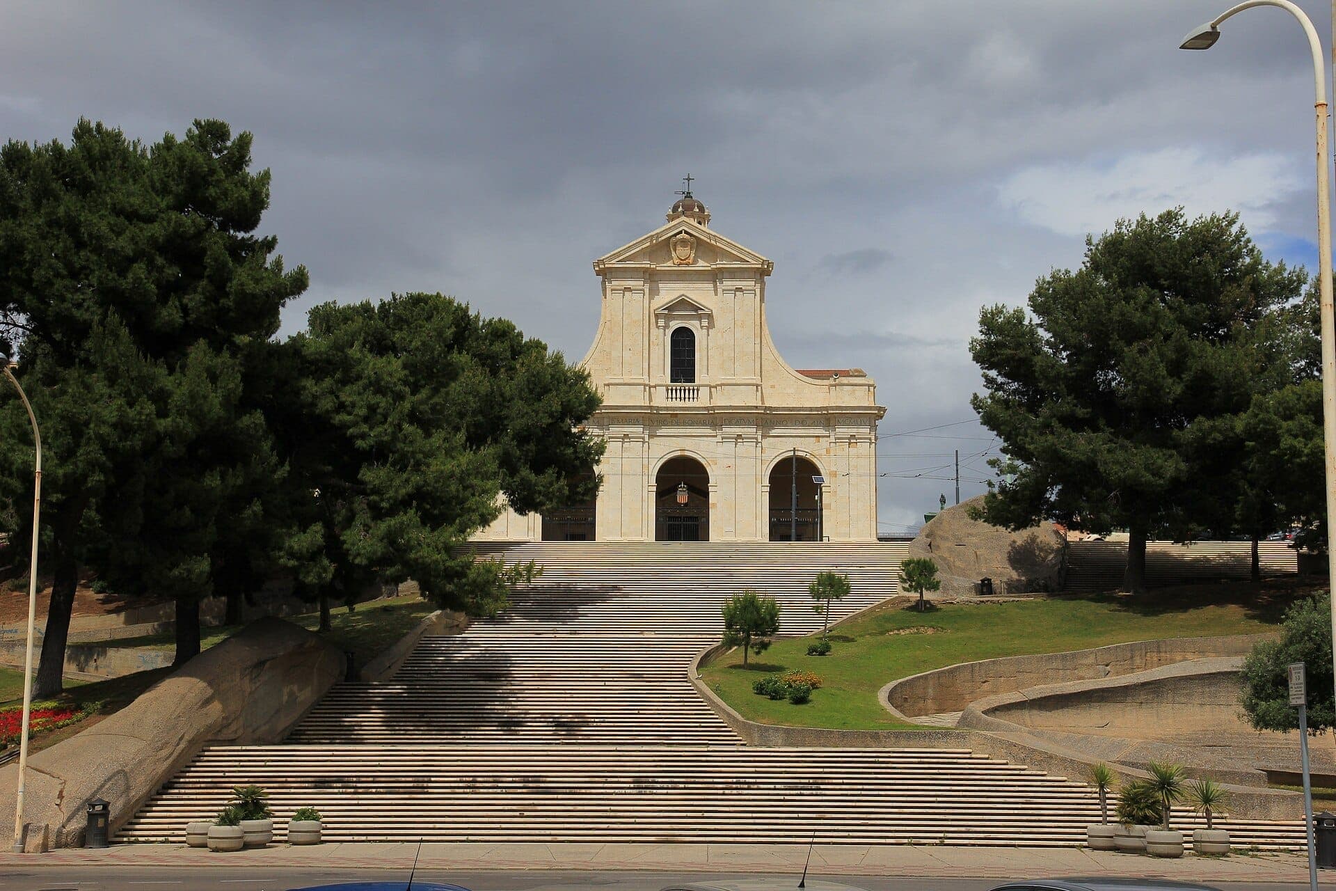 Santuario di Nostra Signora di Bonaria con su gran escalinata, rodeado de árboles verdes y un dramático cielo nublado en Cagliari.