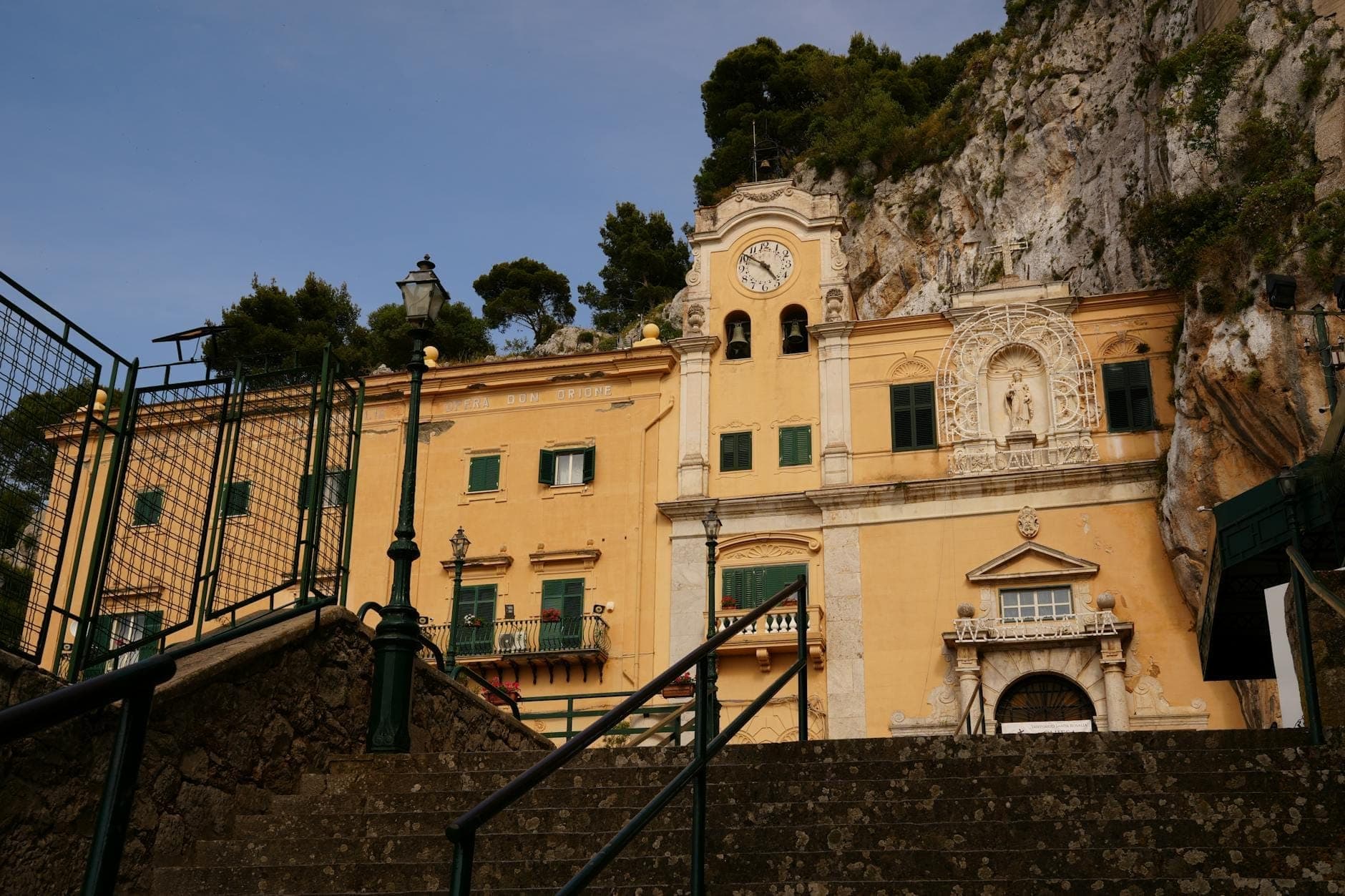 Tangga menuju Sanctuary of Santa Rosalia yang dibangun di tebing batu Monte Pellegrino di luar Palermo, dengan fasad berwarna kuning, menara jam, dan pemandangan alam sekitar yang indah.