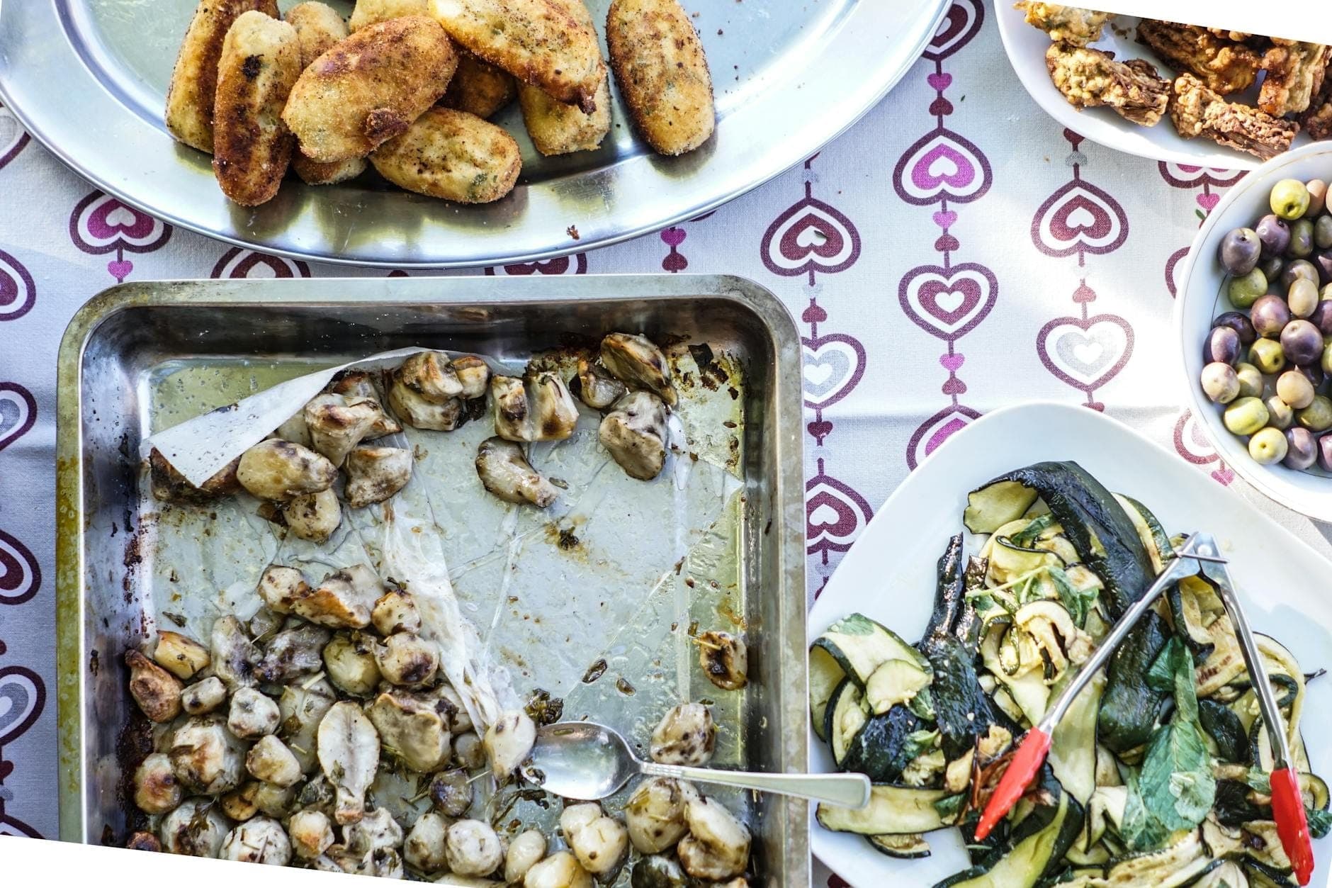 Bandejas de comida casera y rústica que incluyen patatas asadas, ñoquis, verduras a la parrilla, aceitunas y antipasti sobre un mantel estampado, evocando una comida de granja italiana.