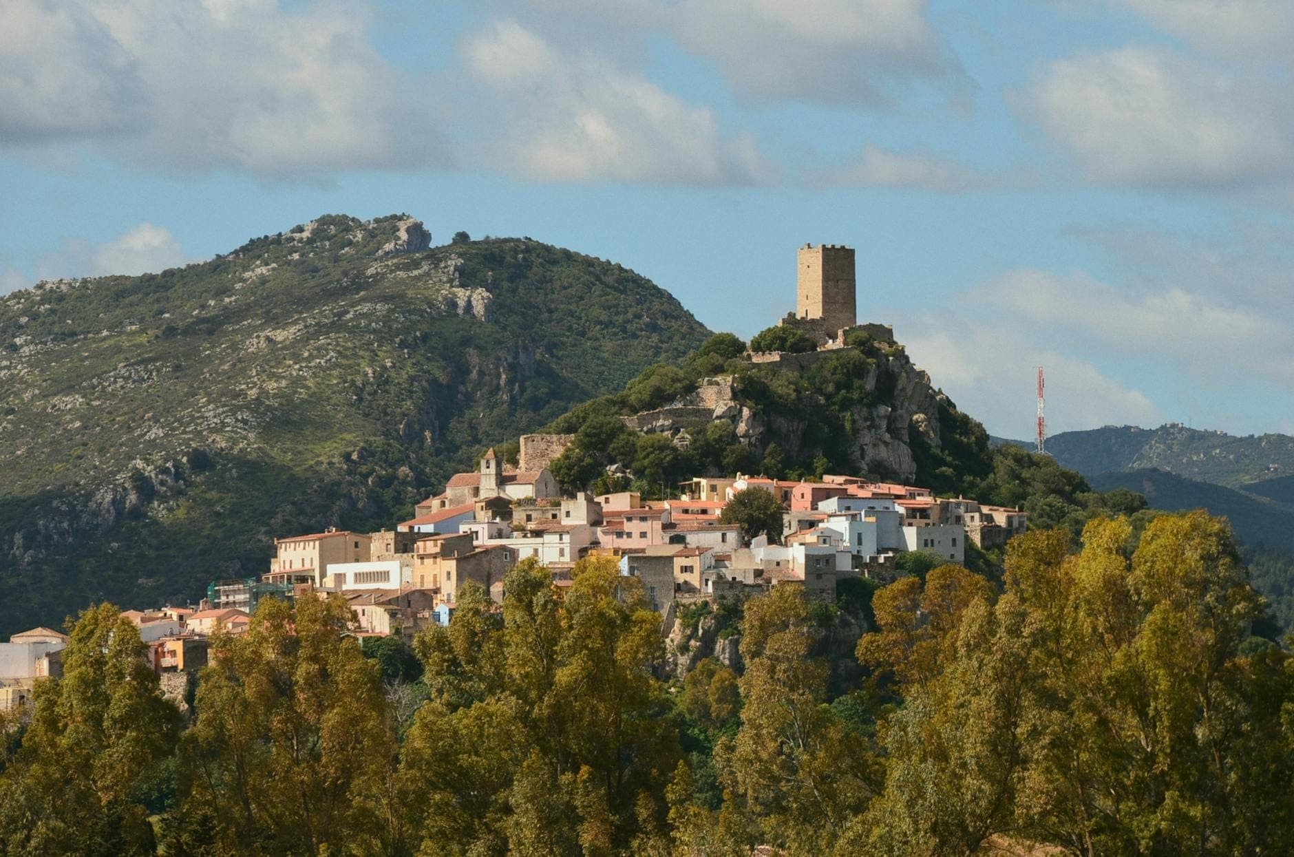 Pueblo sardo en una colina con casas agrupadas y una torre de piedra histórica con vistas a colinas boscosas verdes y cielos parcialmente nublados.