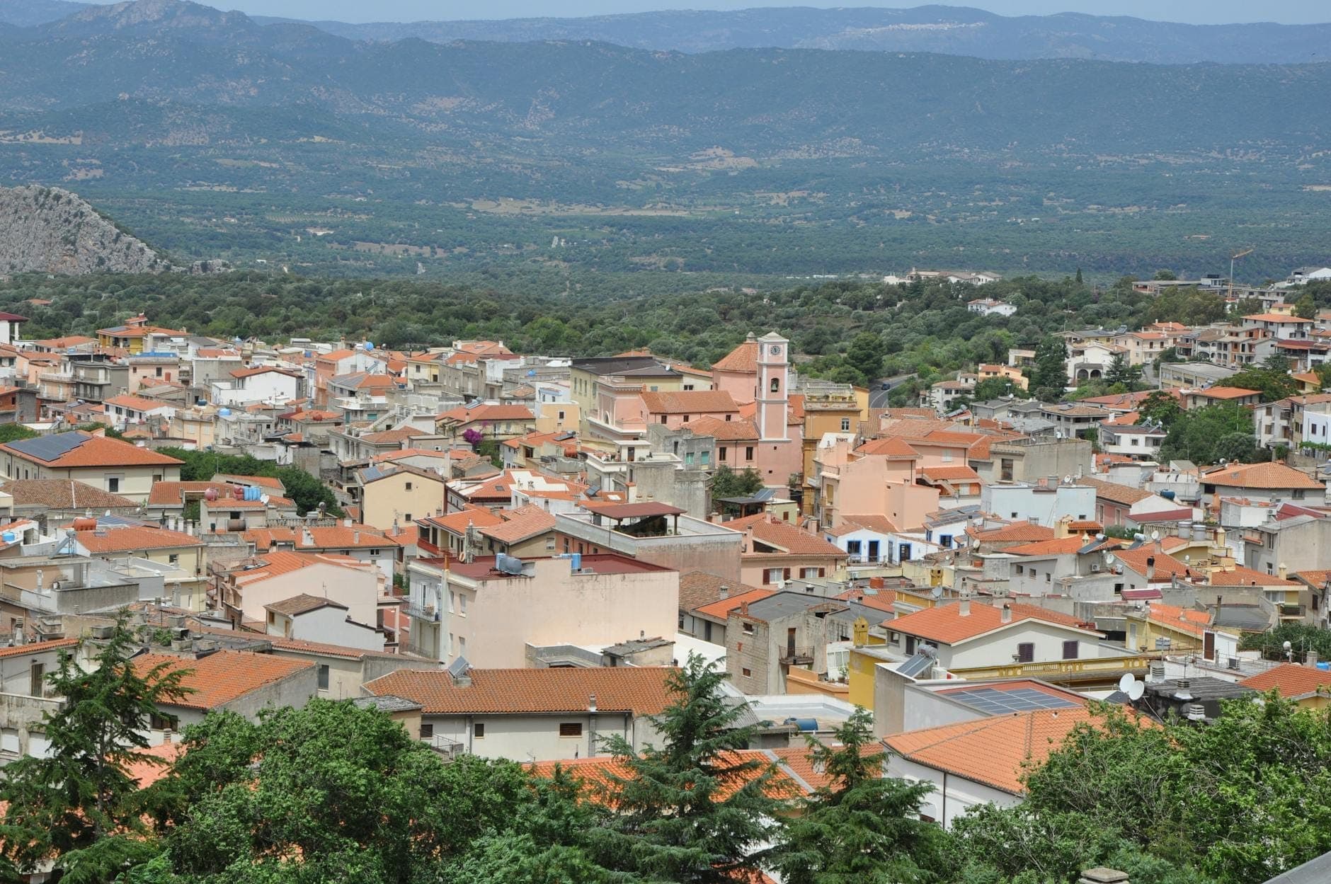 Vista panorámica de un pueblo sardo con tejados de tejas rojas, un campanario y montañas circundantes bajo un cielo despejado.