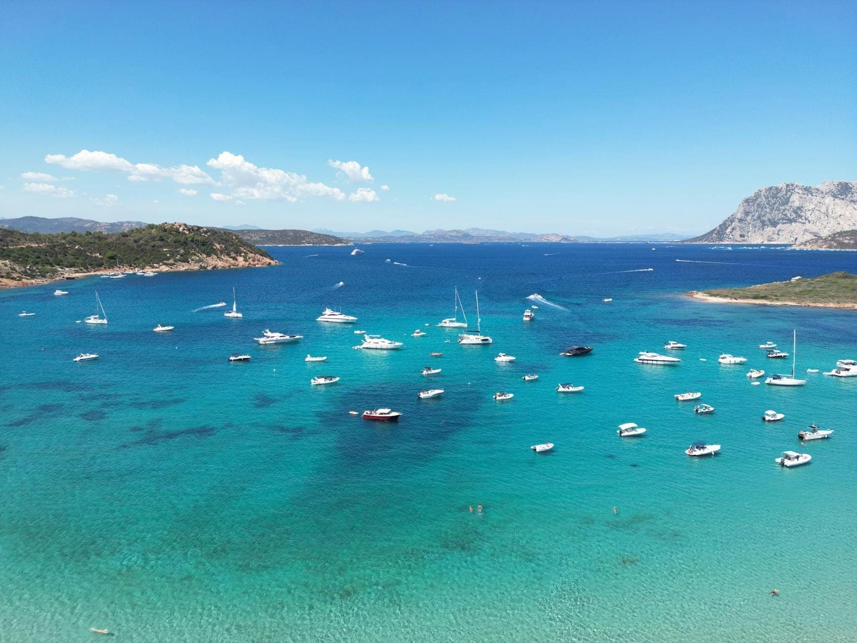 Vista aérea de aguas turquesas salpicadas de numerosas embarcaciones ancladas cerca de las pintorescas islas de Cerdeña, con montañas y cielo despejado al fondo.
