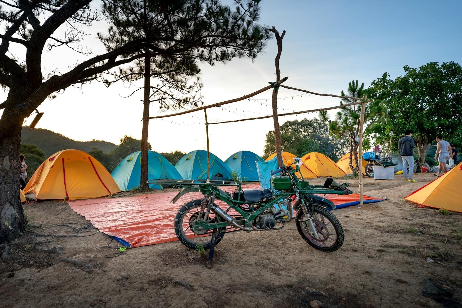 Coloridas tiendas de campaña bajo pinos en un campamento con vistas espectaculares, con una moto y personas al fondo, evocando la aventura de acampar en Cerdeña.