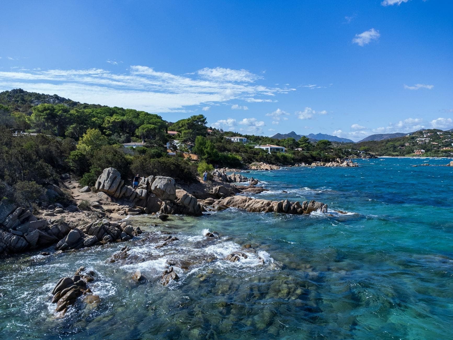 Costa rocosa de Cerdeña con aguas turquesas cristalinas, pinos y zonas urbanizadas al fondo bajo el sol, ideal para acampar cerca de las principales atracciones.