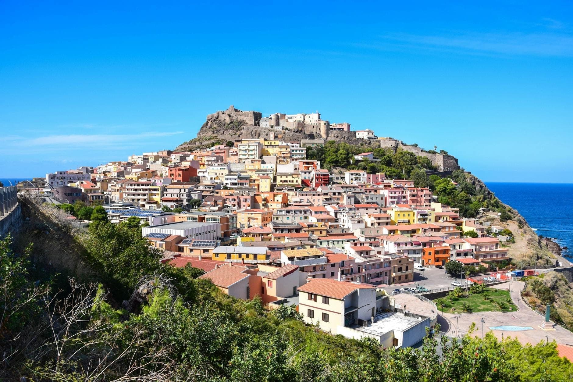 Colorful hillside town with a medieval castle on top, overlooking the sea under a clear blue sky in Sardinia, Italy.