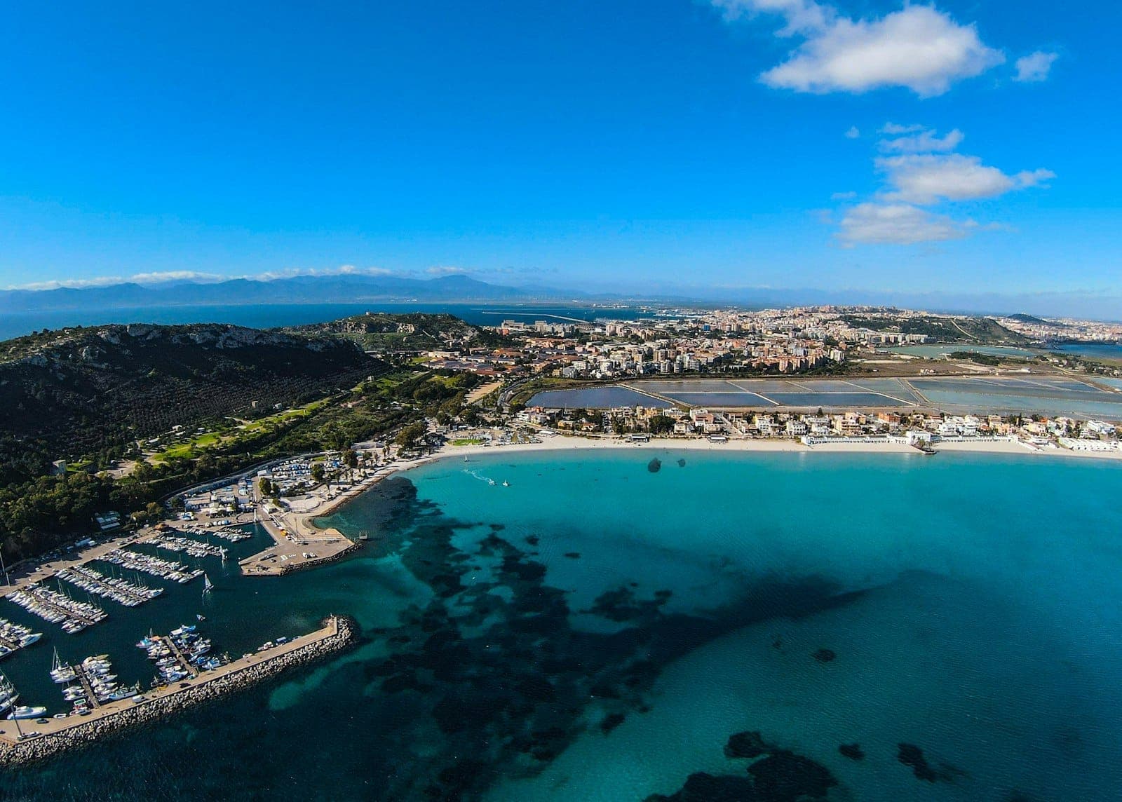Vista aérea de la costa de Cagliari en Cerdeña con aguas azules cristalinas, marina, edificios de la ciudad y montañas lejanas bajo un cielo despejado.