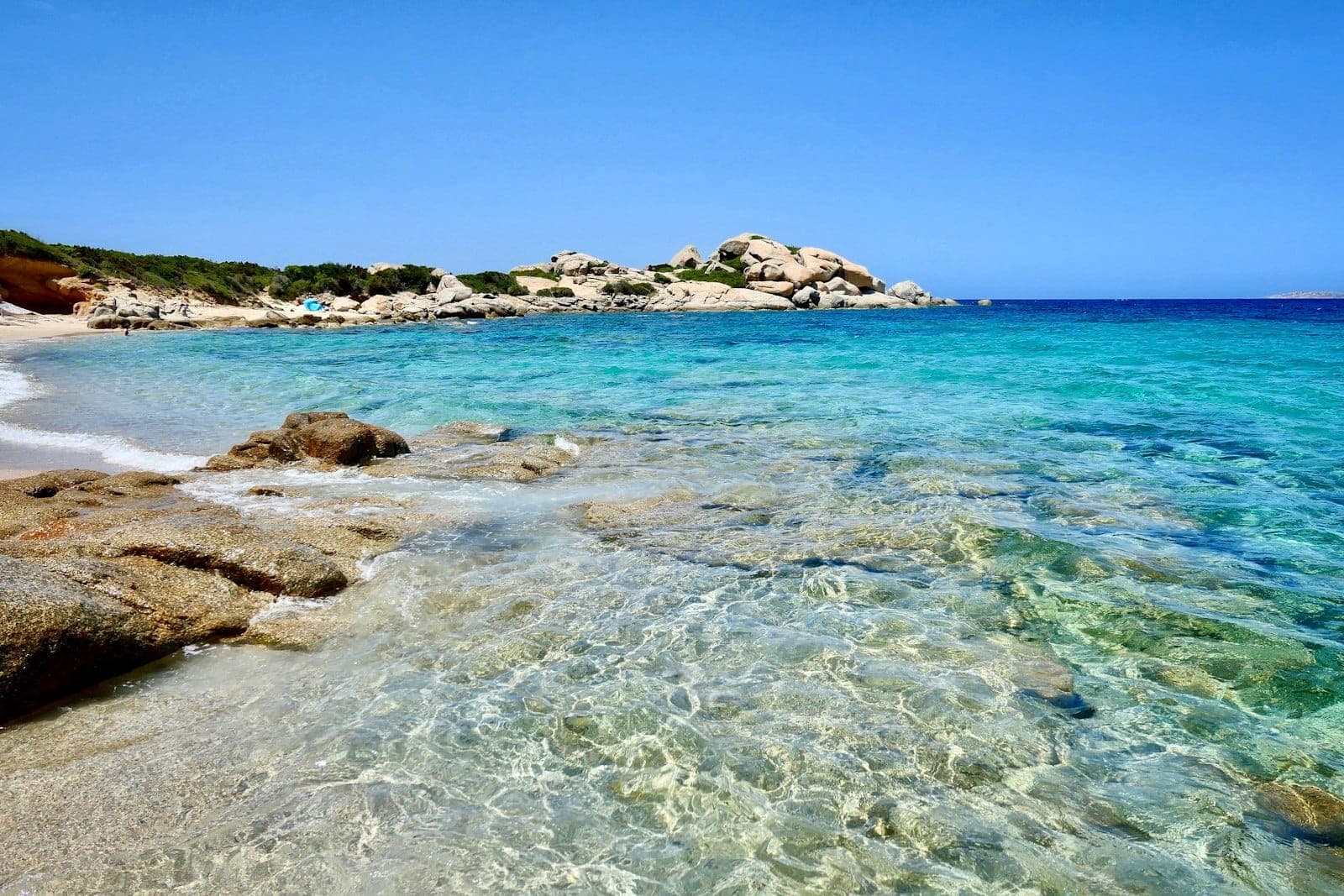 Agua turquesa cristalina y playa de arena blanca con rocas de granito en la costa de Cerdeña bajo un cielo azul brillante.