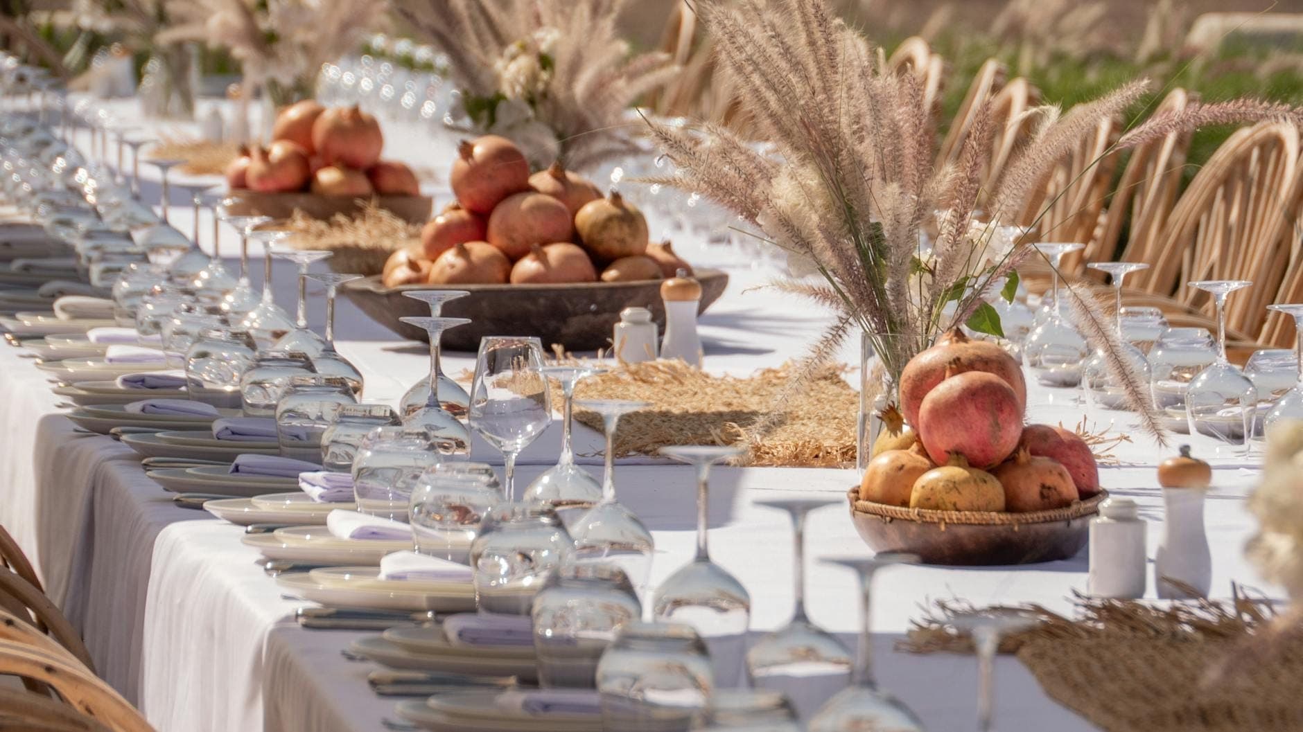 Larga mesa de granja al aire libre preparada para una comida con manteles blancos, platos, vasos, centros de frutas y decoraciones rústicas en un entorno soleado.