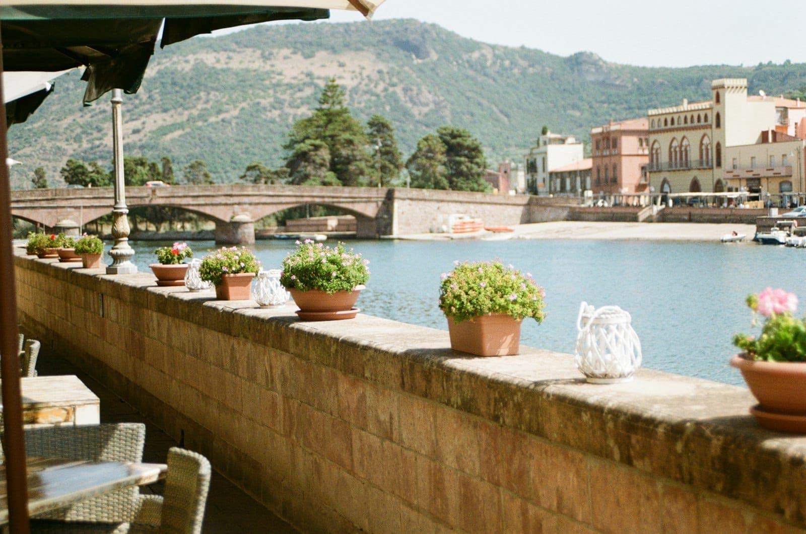 Terraza de un café con vistas al agua en Cerdeña, con plantas en macetas, mesas y sillas, frente a un río y edificios históricos bajo un cielo soleado.