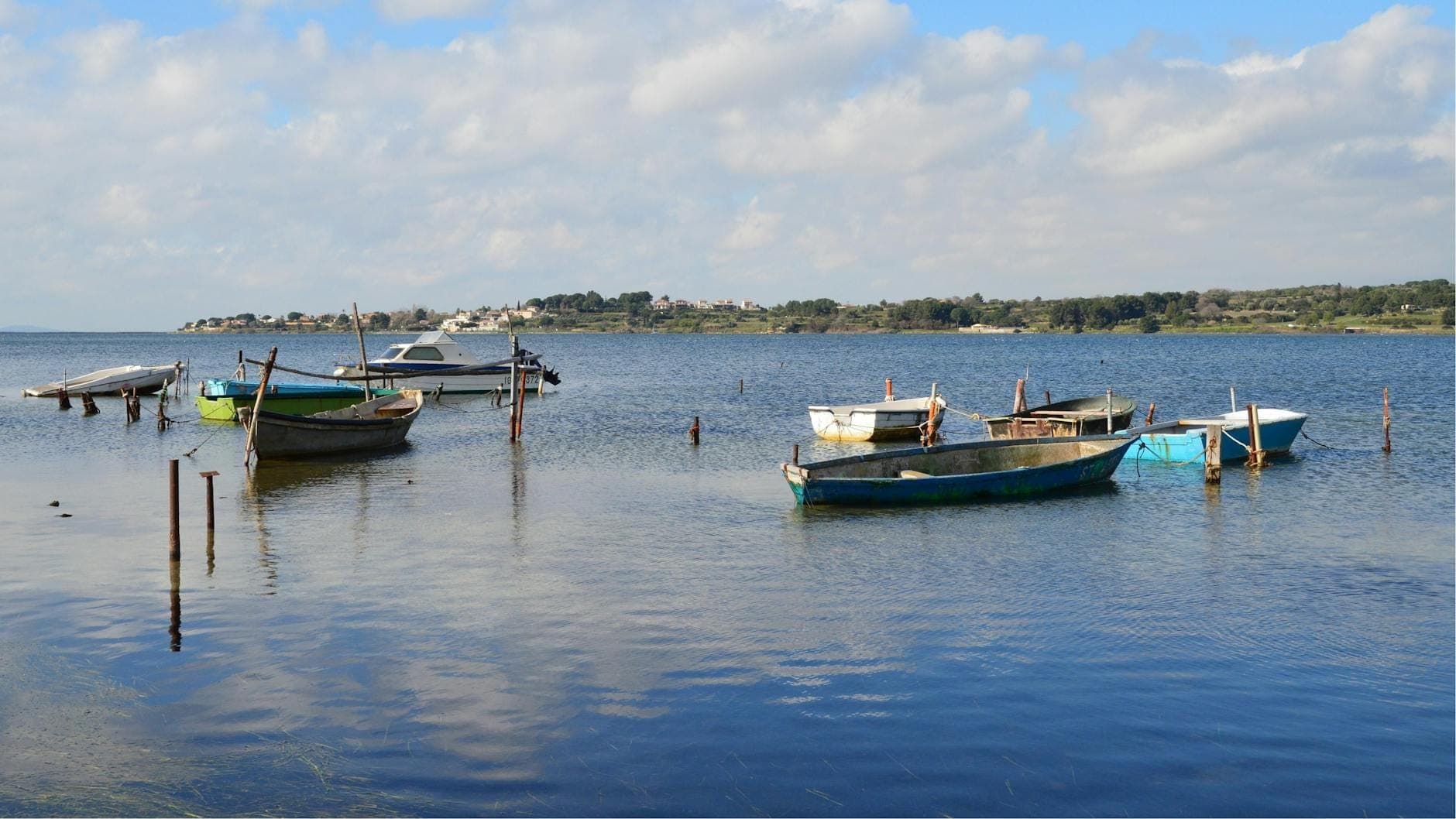 Pequeñas barcas de pesca ancladas en una laguna tranquila con colinas lejanas y un cielo parcialmente nublado, evocador de la pesca costera de Cerdeña.