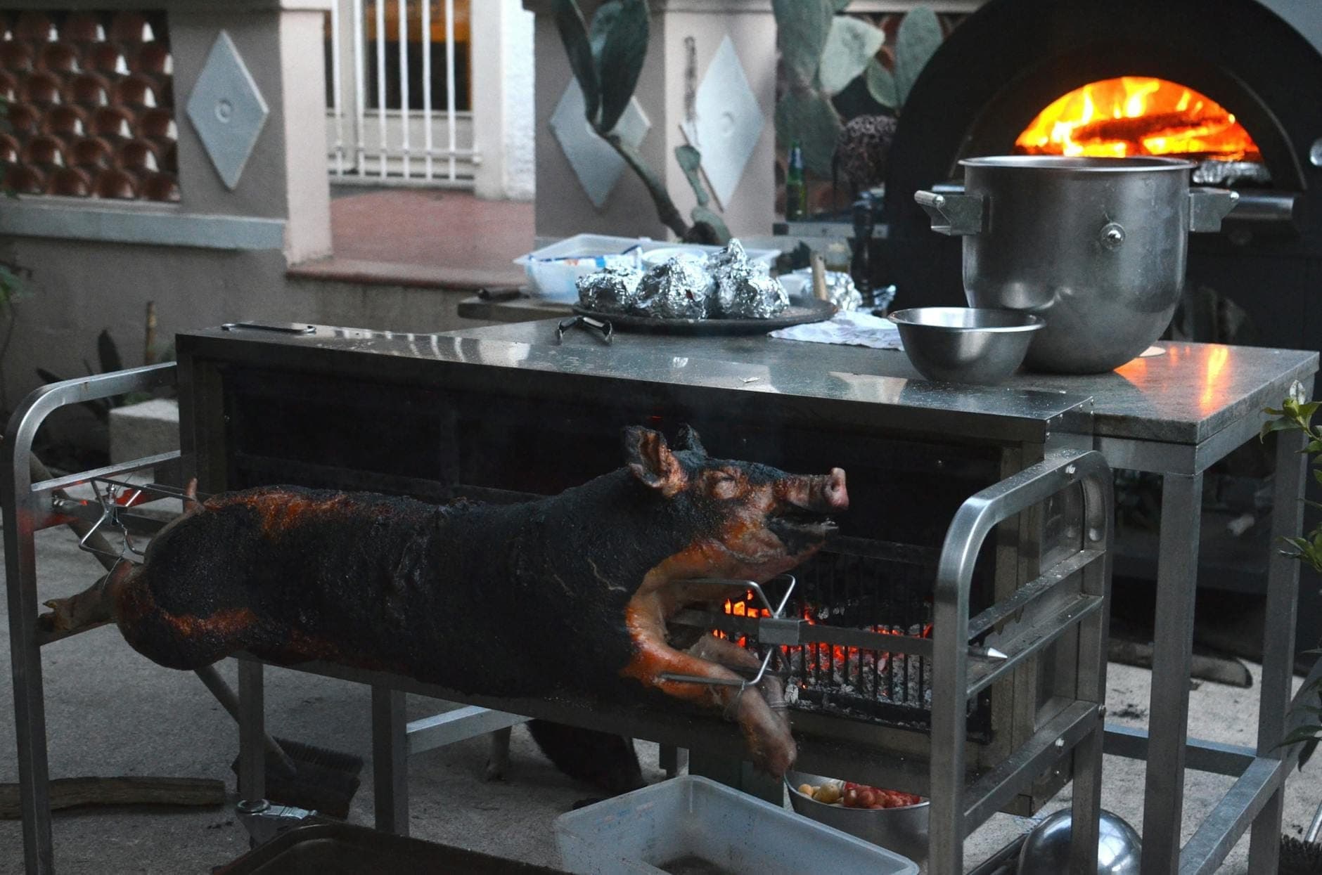Cochinillo entero asándose en un espeto sobre brasas al aire libre con horno de fuego al fondo. Escena tradicional de preparación de carne.
