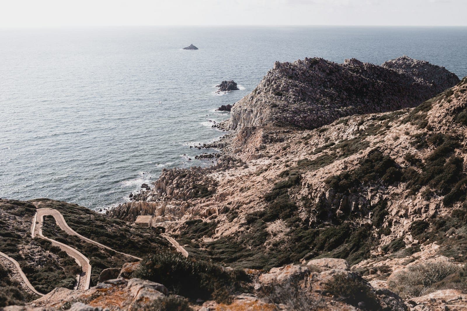 Una costa escarpada de Cerdeña con senderos sinuosos, acantilados rocosos y vegetación escasa que se asoma al tranquilo mar Mediterráneo bajo un cielo suave.