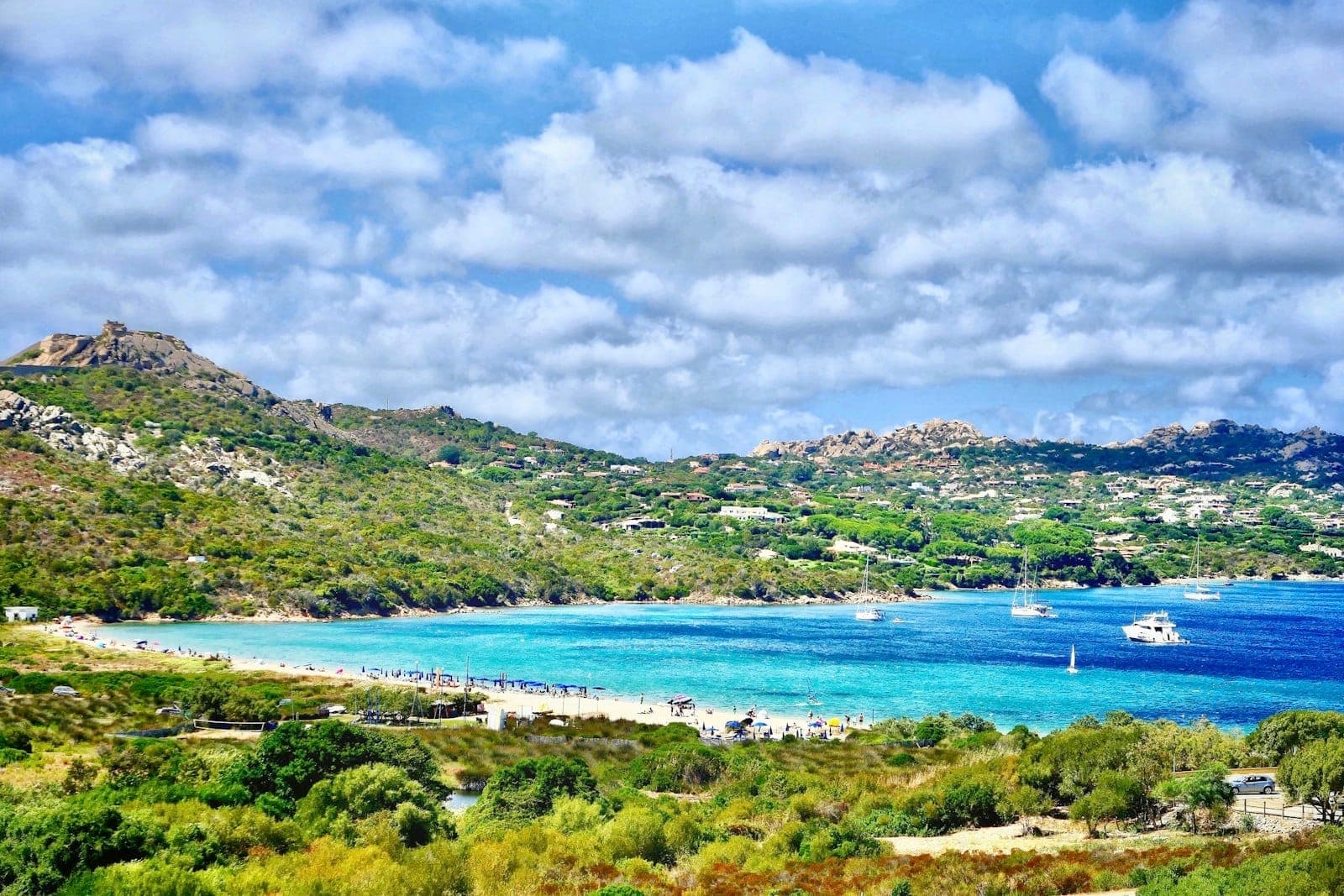 Bahía sarda soleada con agua turquesa, colinas verdes, playa de arena y barcas dispersas, bajo un cielo con nubes dispersas a finales de primavera.