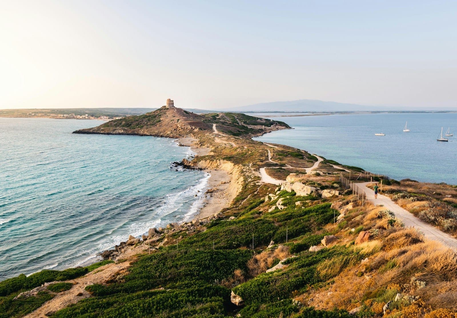 Vista panorámica de la costa de Cerdeña con aguas turquesas, acantilados escarpados, barcos fondeados frente a la orilla, senderos serpenteantes y una torre histórica bañada por el sol a lo lejos.