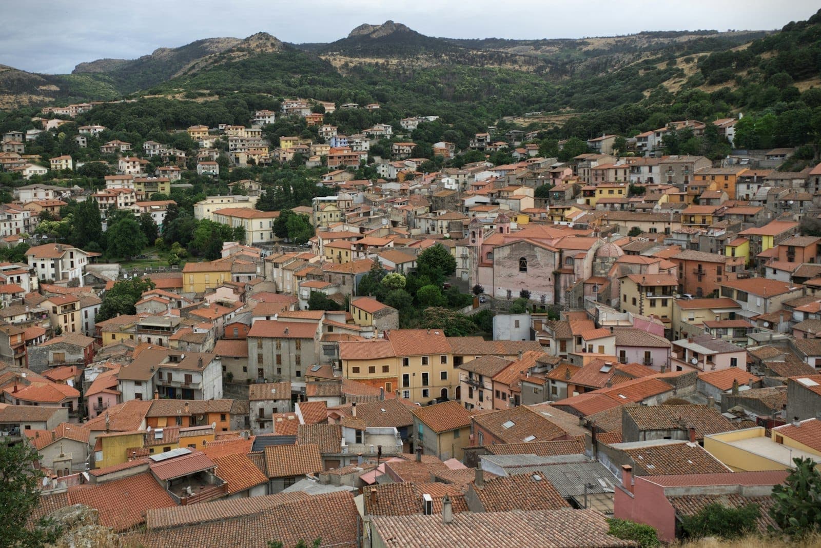Vista panorámica de un pueblo de montaña sardo con tejados de terracota y colinas verdes circundantes, típico de los pueblos de Barbagia que acogen el festival.